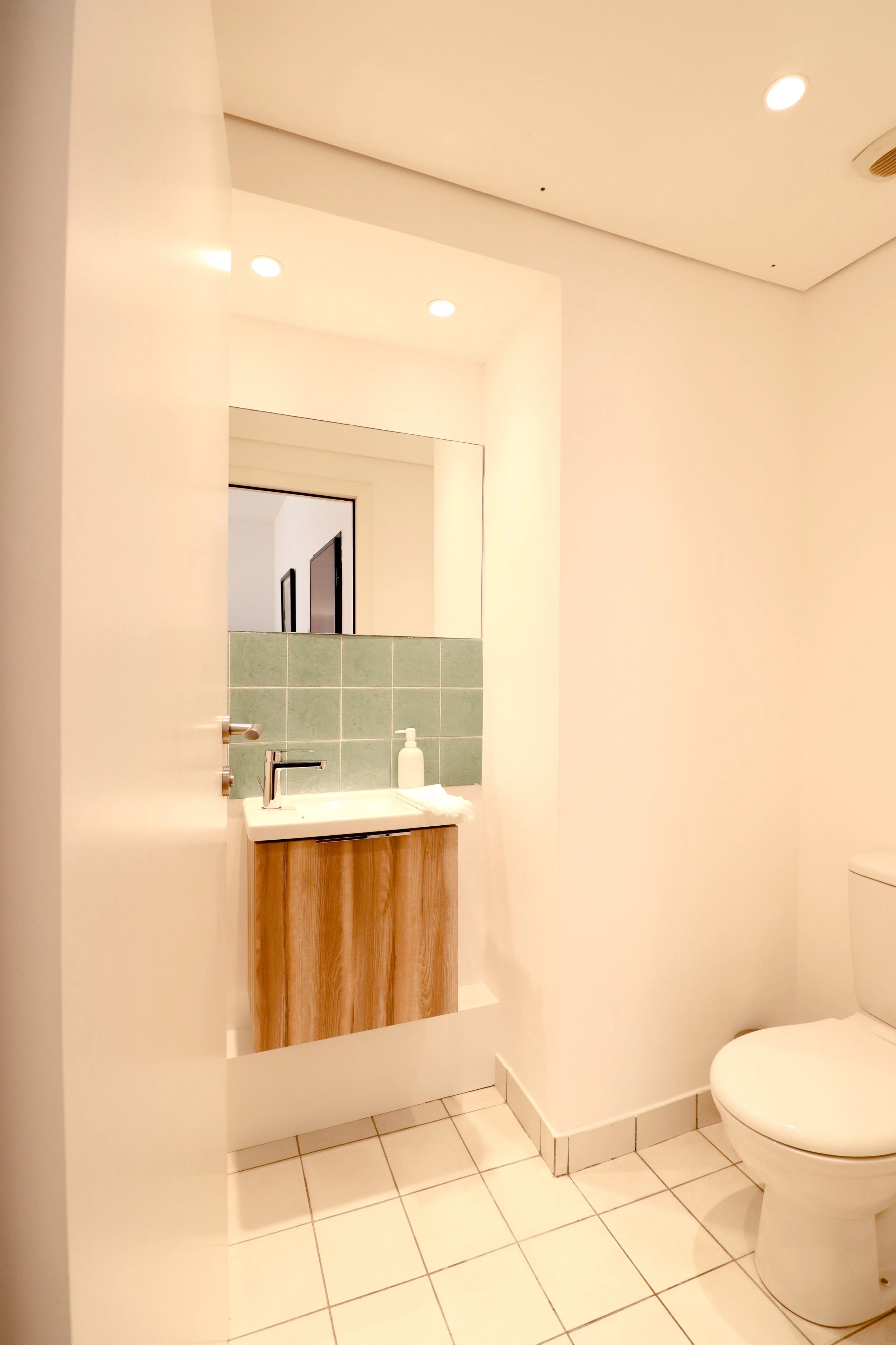 Intérieur d'une salle de bain avec un miroir, un lavabo en bois, un distributeur de savon blanc, un WC blanc, des carreaux de sol blanc avec des lignes grises, et un plafond avec plusieurs lumières encastrées.