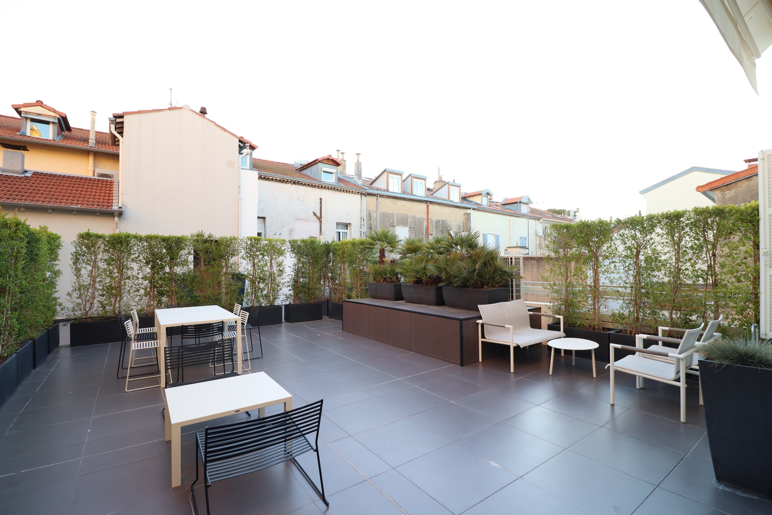 Vue d'une terrasse extérieure avec des meubles en blanc et noir, des plantes en pots et une barrière végétale, surplombant des maisons avec toits en tuiles rouges