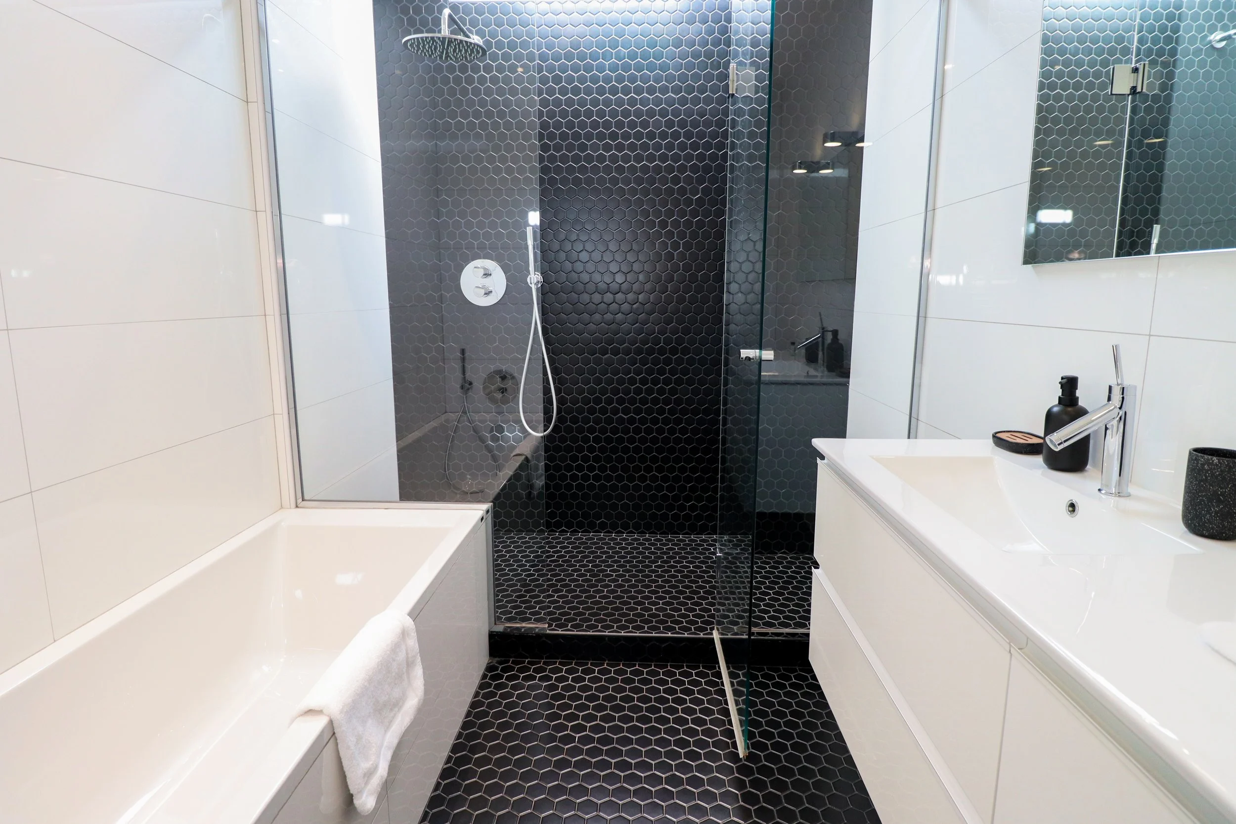 Une salle de bain moderne avec une cabine de douche en verre avec un mur en carreaux noirs hexagonaux, une baignoire blanche, un lavabo blanc avec des accessoires noirs, et un miroir.