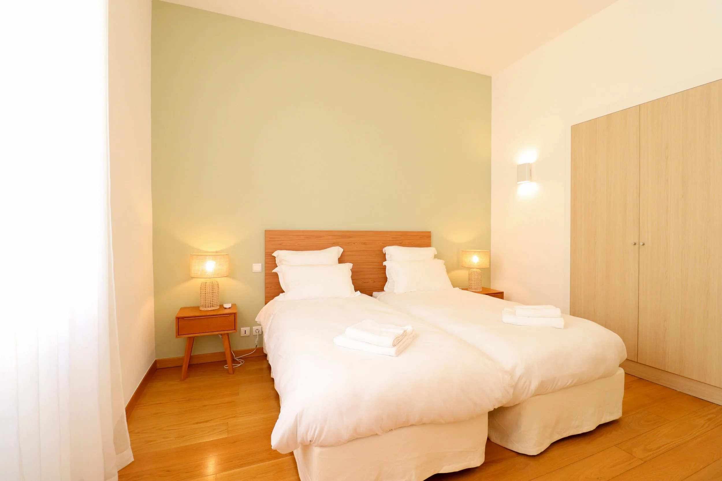 Chambre avec deux lits doubles en bois clair, lampes de lecture déco, petite table de nuit, armoires en bois clair, et linge plié sur les lits.
