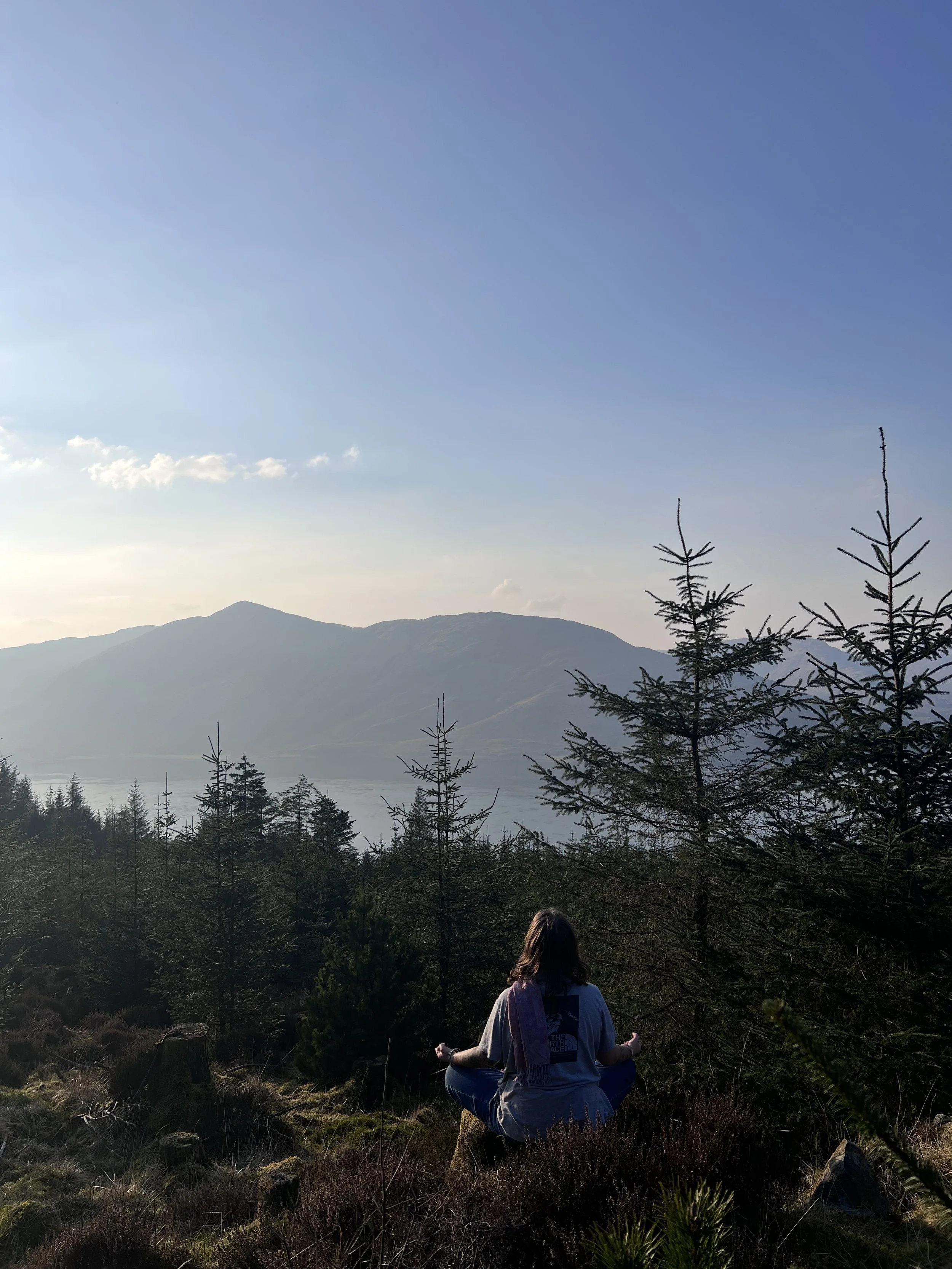 Person meditating in a forest overlooking a mountain and lake under a clear blue sky.