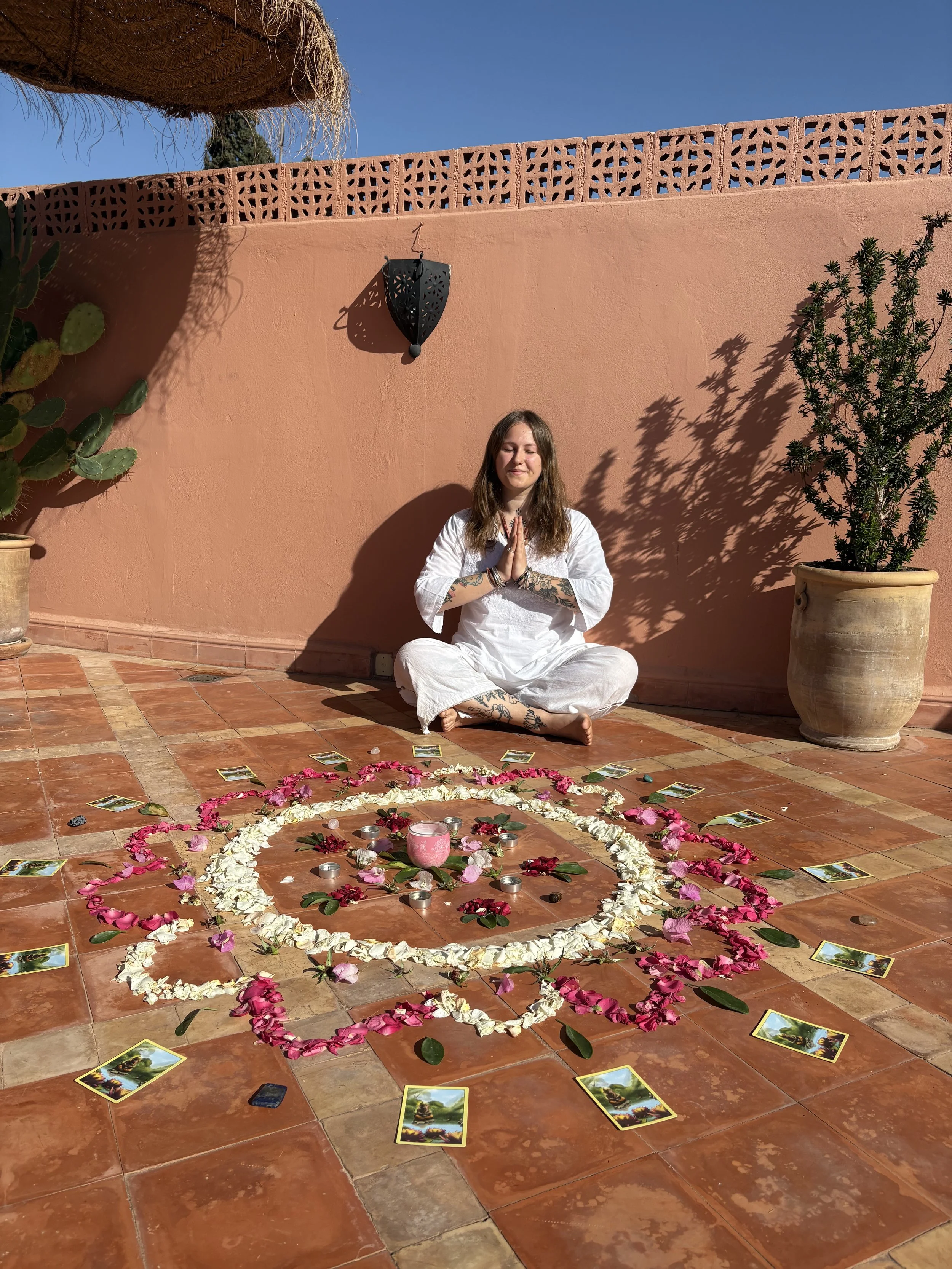 A cozy yoga or meditation room with mats arranged in a circle around a stone centerpiece with candles and a small statue. Petals are scattered across the floor. Large windows reveal a view of a green forest outside. A lit fireplace is in the corner of the room.