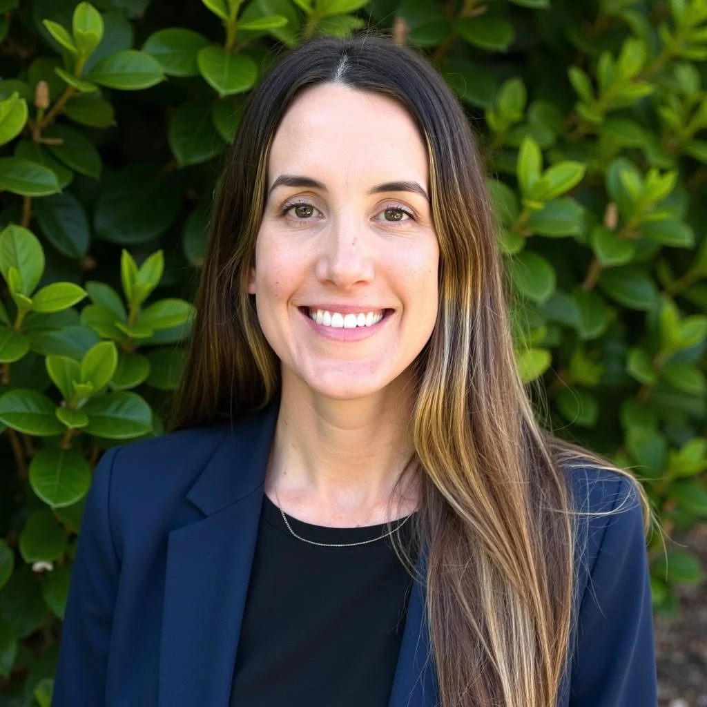 A woman with long brown hair, fair skin, and a bright smile standing outdoors in front of green leafy bushes, wearing a dark navy blazer and a black top.
