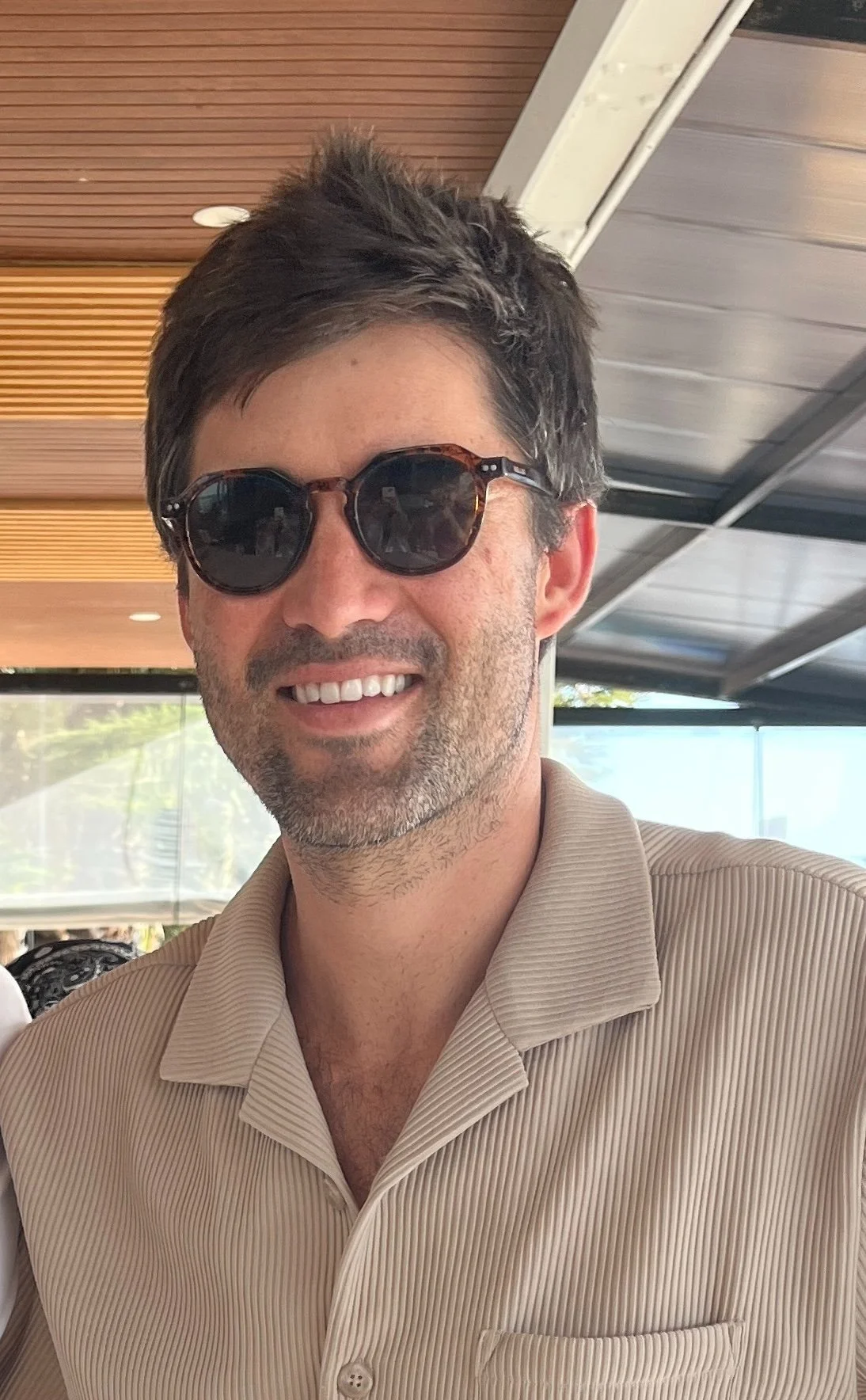 A man with dark hair, sunglasses, and a light tan collared shirt smiling in an outdoor or semi-outdoor setting.