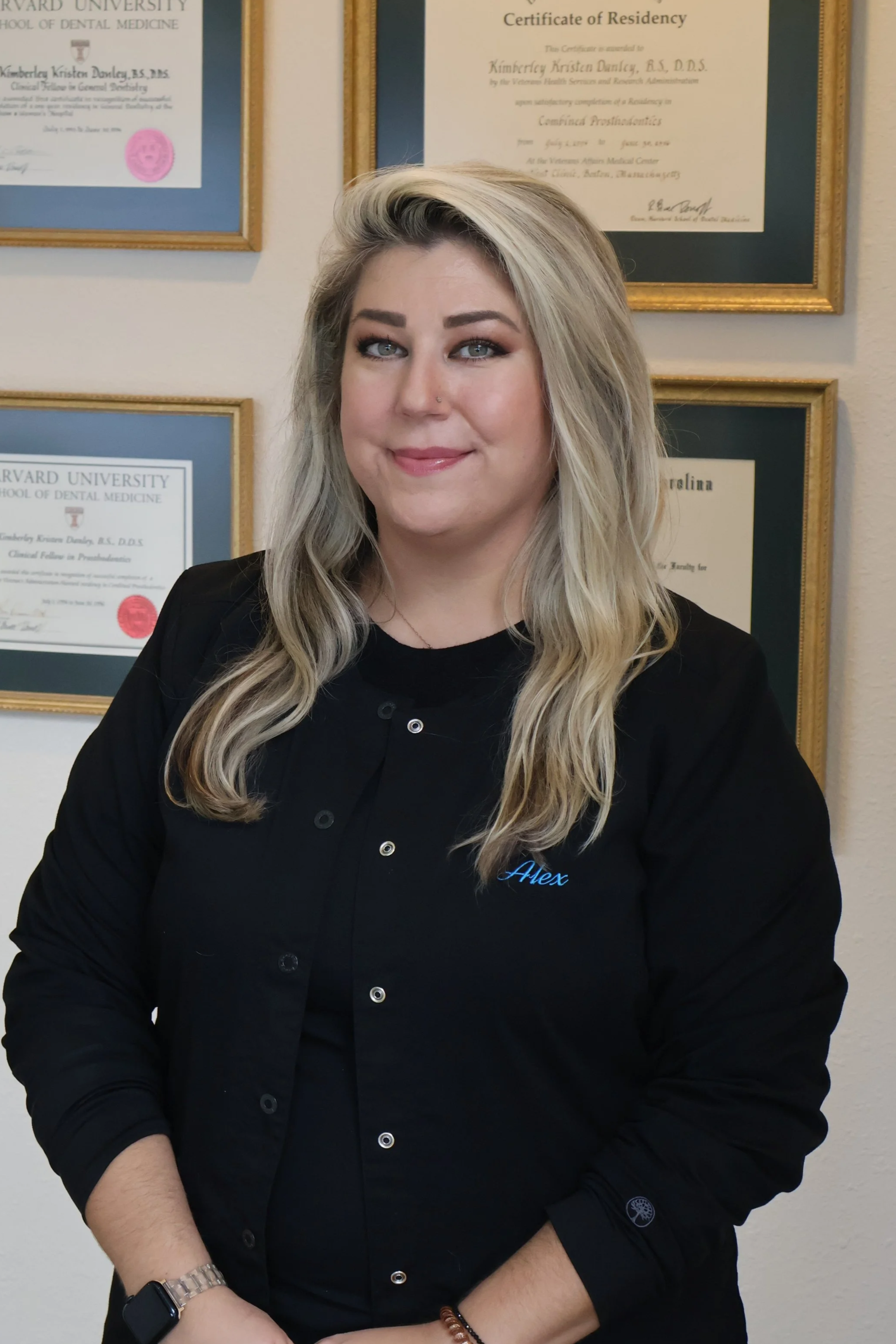 A woman with long, curly blonde hair smiling at the camera. She is wearing a black shirt embroidered with the name 'Alex' and a necklace. In the background, there are framed diplomas and certificates on a wall.
