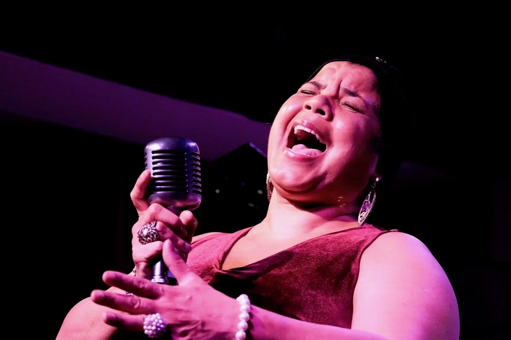 A woman singing passionately with closed eyes, holding a vintage microphone, wearing earrings, rings, and a sleeveless top on stage.