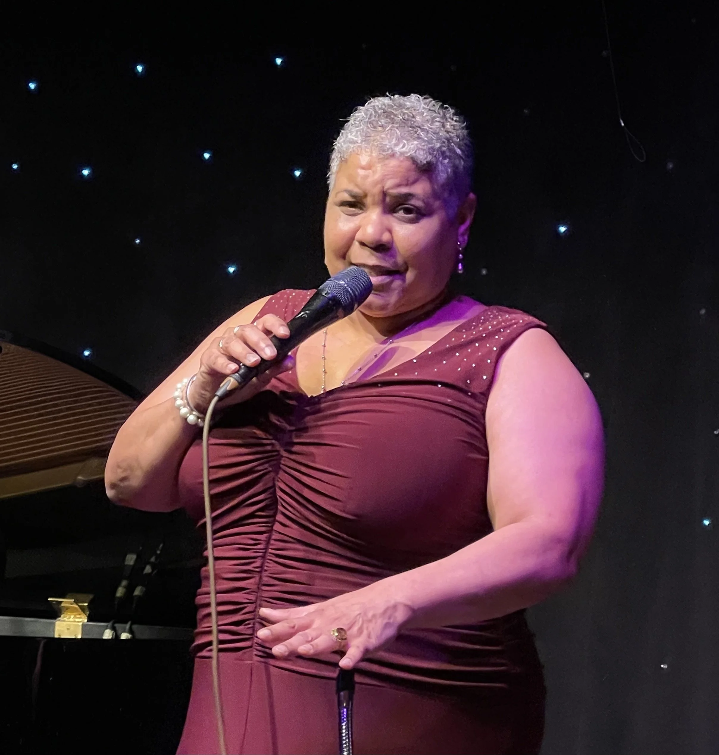 A woman with short, curly gray hair singing into a microphone on a stage with a starry backdrop, wearing a maroon dress with rhinestone details.