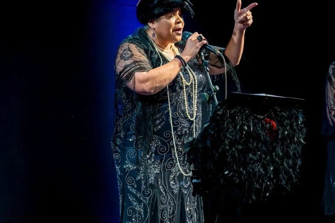 A woman with dark hair and wearing black lace and jewelry, singing into a microphone on stage, with a person with curly hair in the foreground.