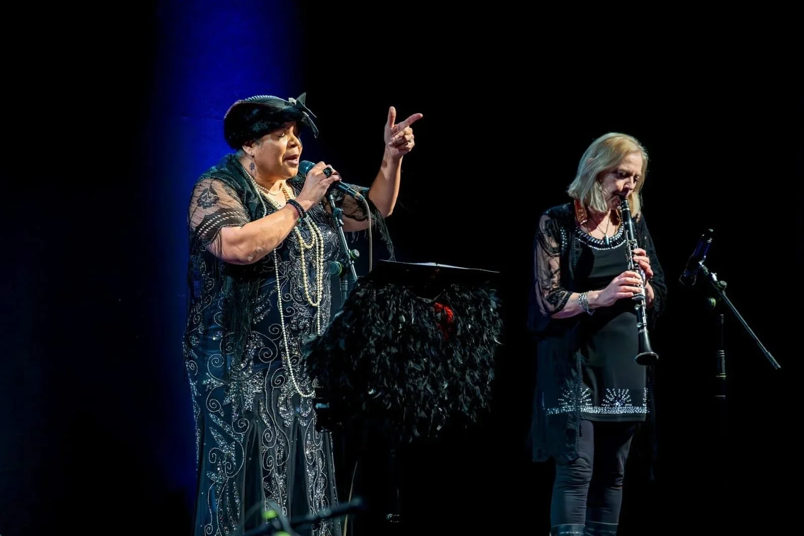 Two women performing on stage: one singing into a microphone, the other playing a clarinet, both dressed in black with decorative details.