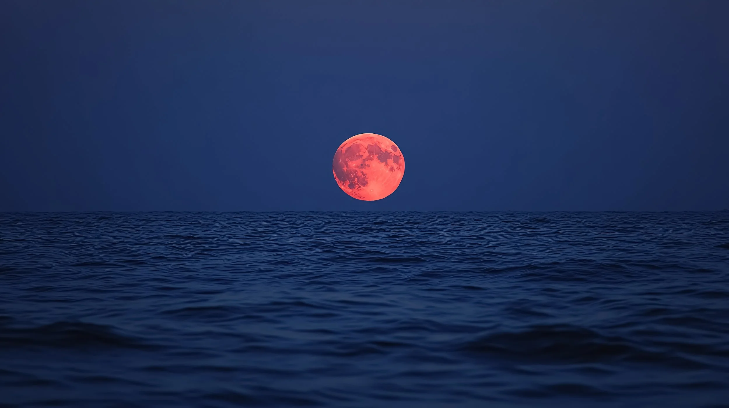 A large, reddish-orange full moon over a dark ocean at night.