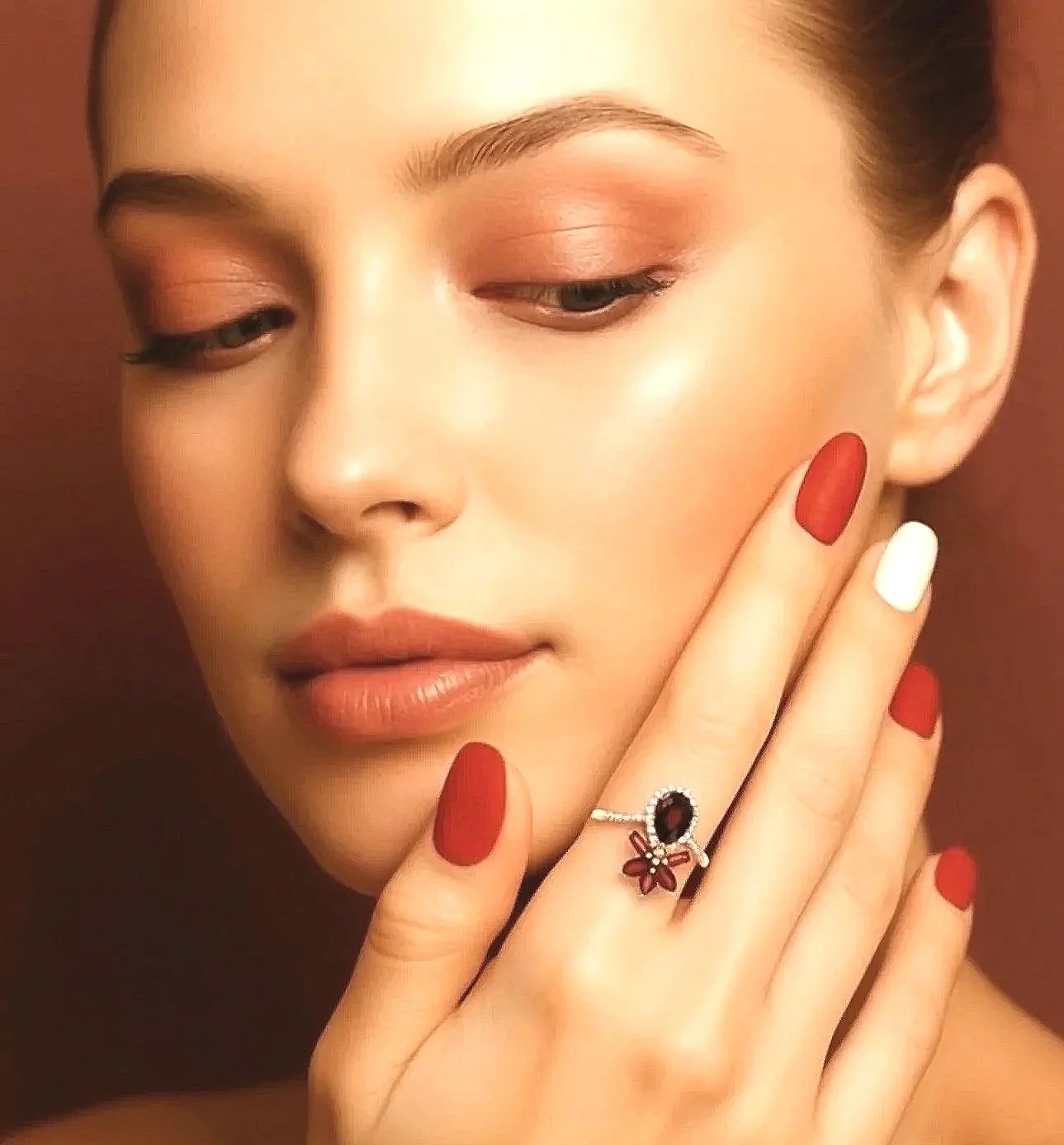 Close-up of a woman with natural makeup, red and white painted nails, and a floral ring on her finger.