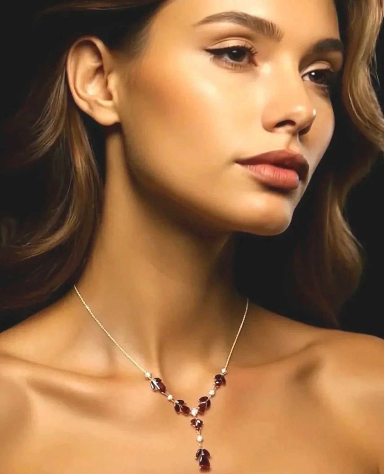 Close-up of a woman with long, wavy brown hair wearing a silver necklace with dark red and clear gemstones.