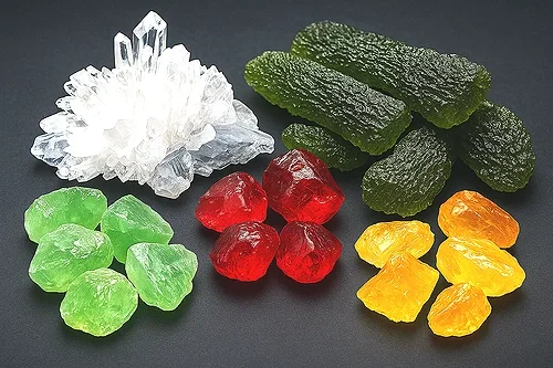 A collection of colorful crystals and mineral stones on a black background, including clear quartz, green jade, red jasper, yellow calcite, and black garnet.