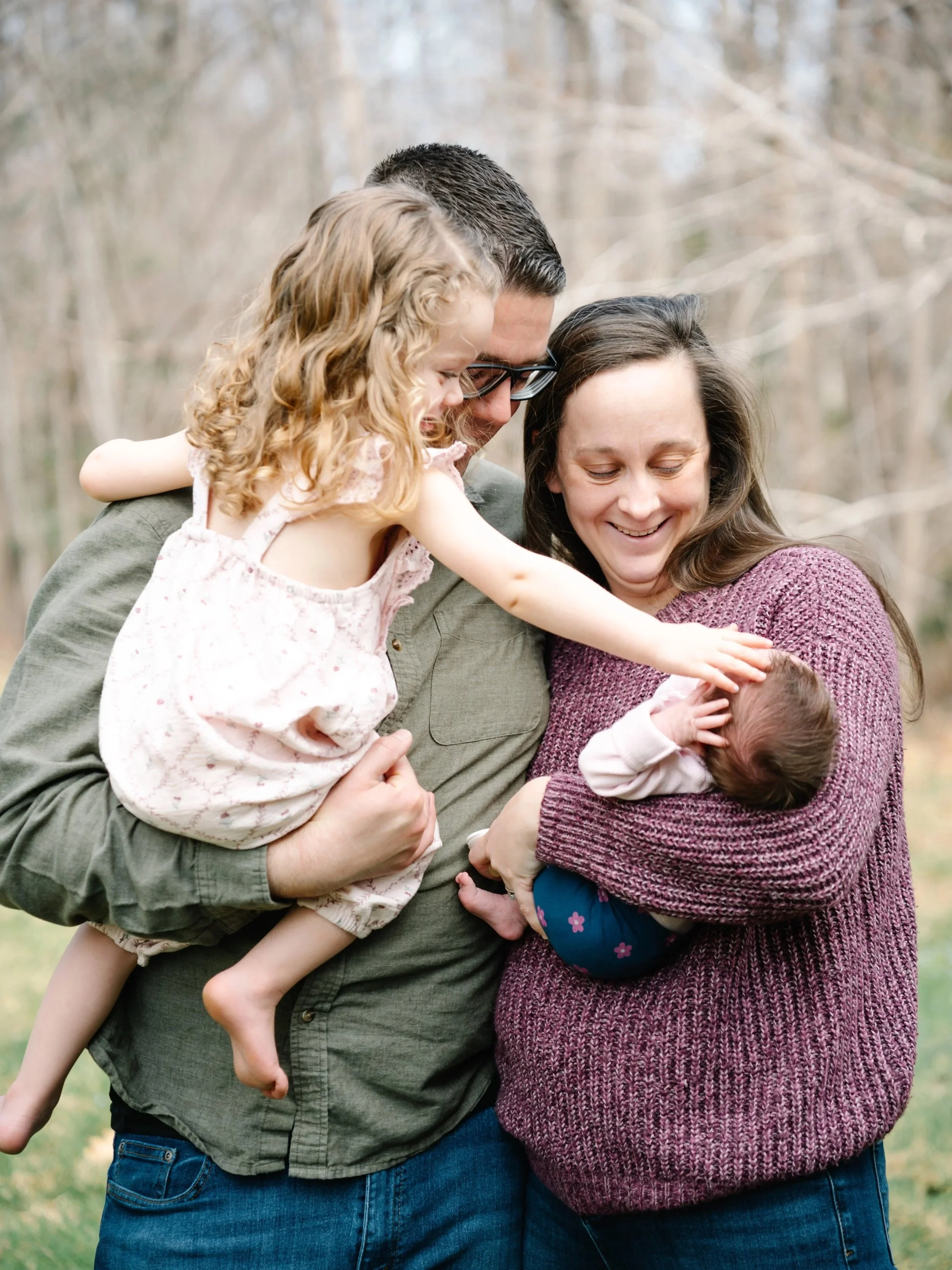 A family of four, consisting of a father, mother, young daughter, and newborn baby, standing together outdoors in a wooded area, smiling and hugging.