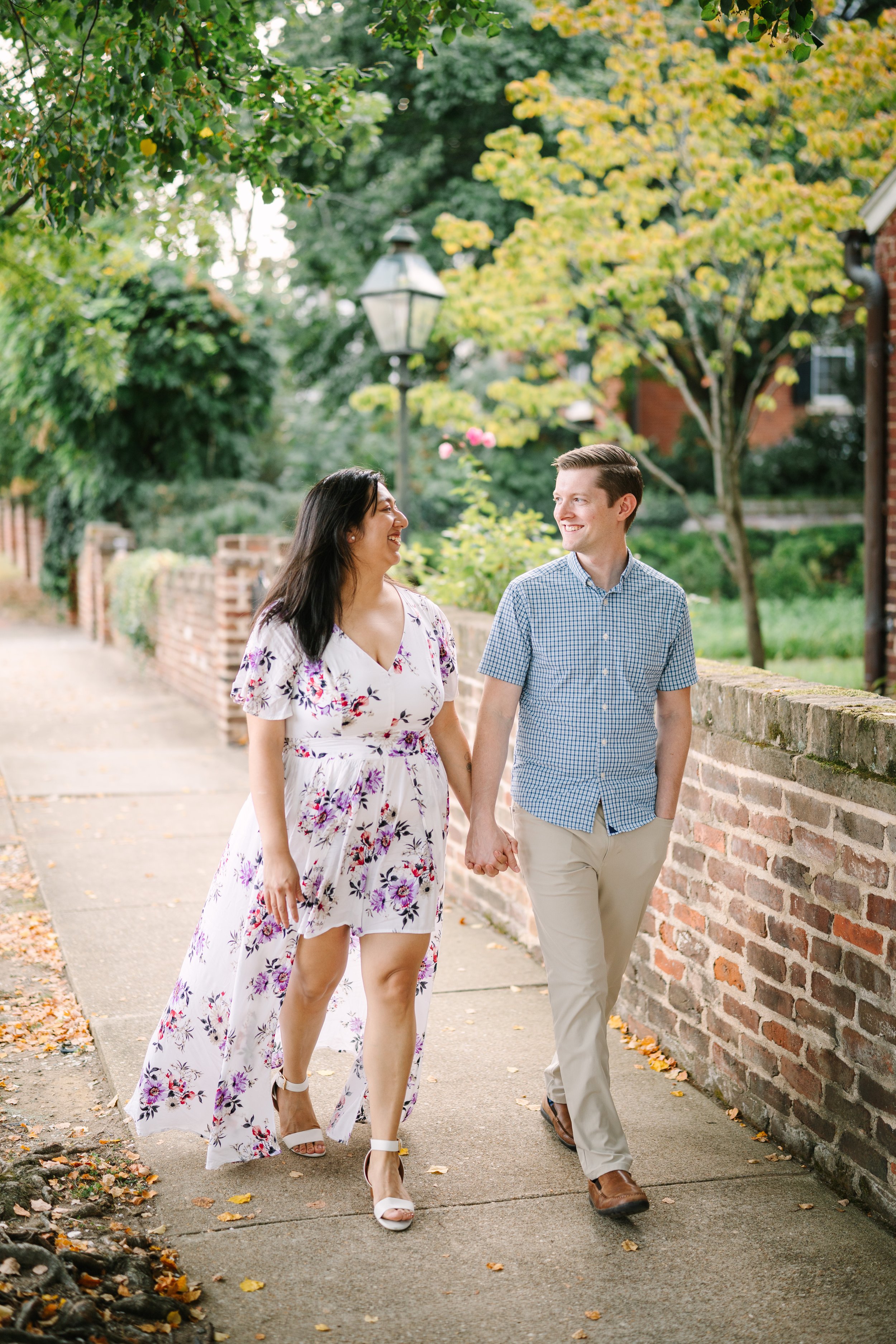 A couple walking hand in hand on a sidewalk in a residential neighborhood with trees and a brick wall, smiling and looking at each other.
