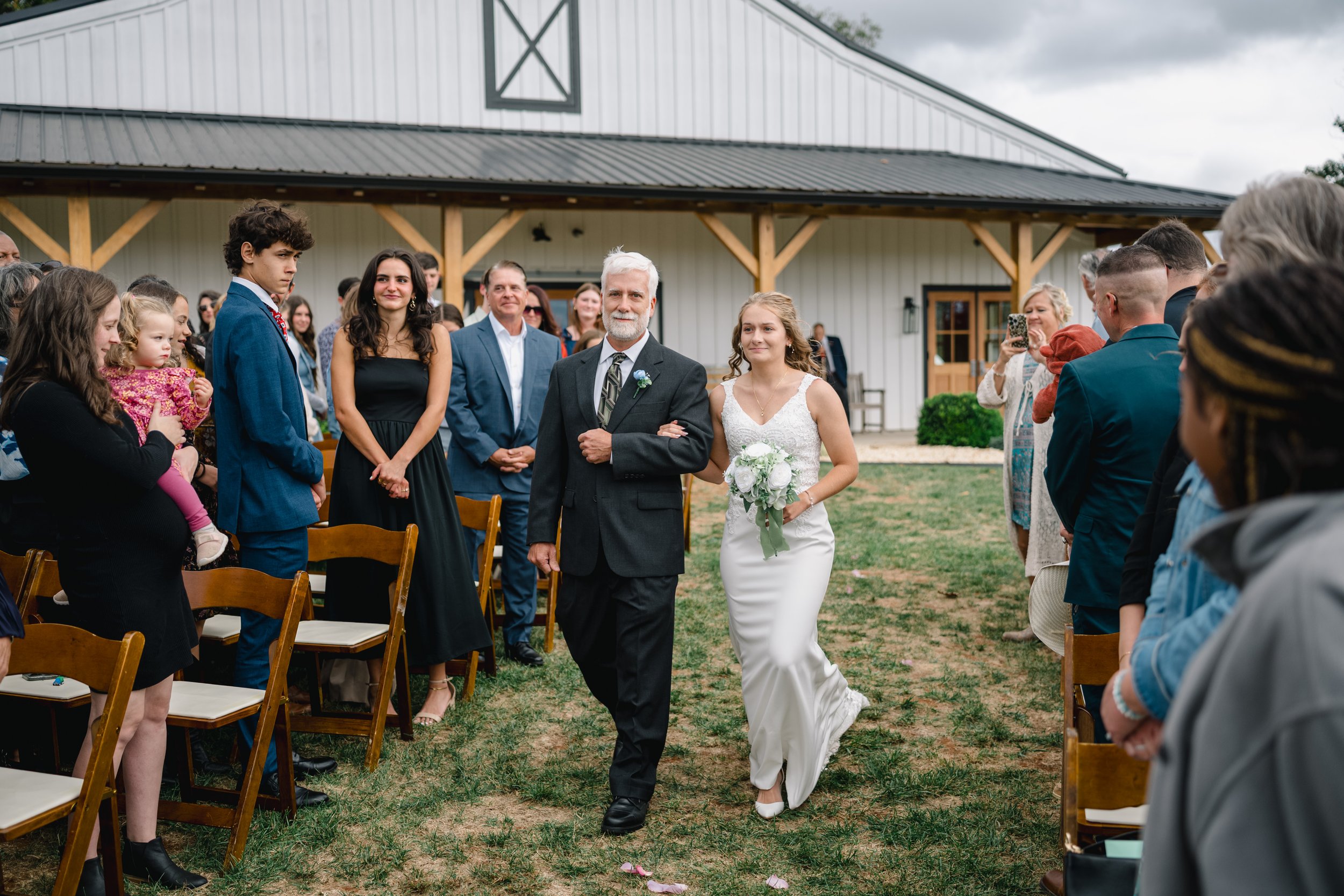 Bride walking down the aisle with her father at an outdoor wedding ceremony in front of a barn, with guests watching and taking photos.