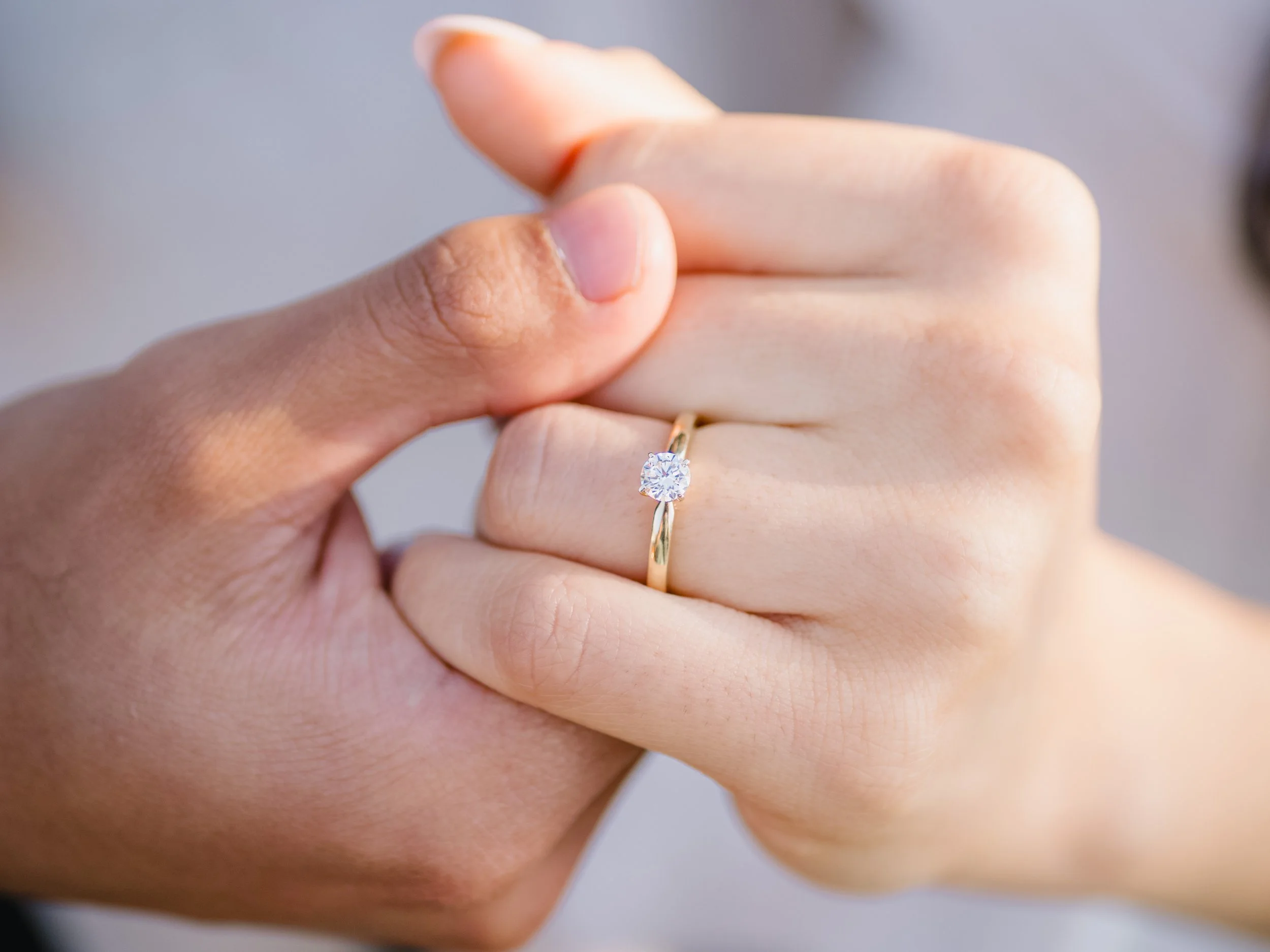 Close-up of a person's hand wearing a gold ring with a large diamond, with another hand gently touching the ring.