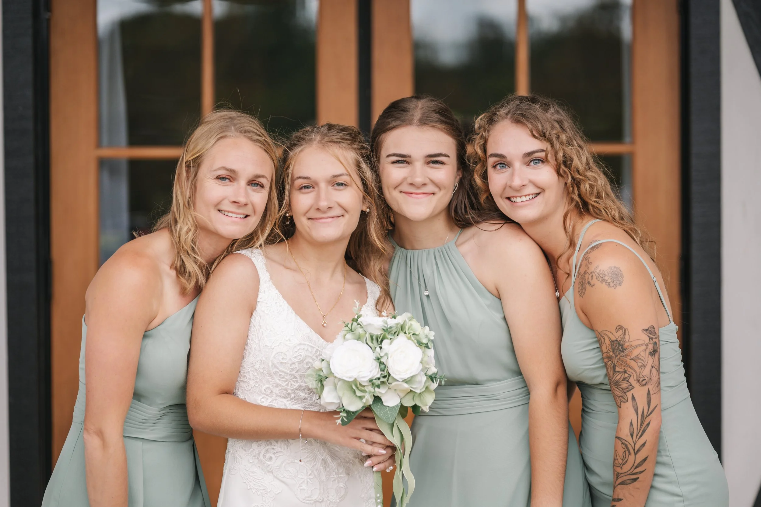 Four women, including a bride holding a bouquet, smiling outside in front of a house with large windows and wooden framing.