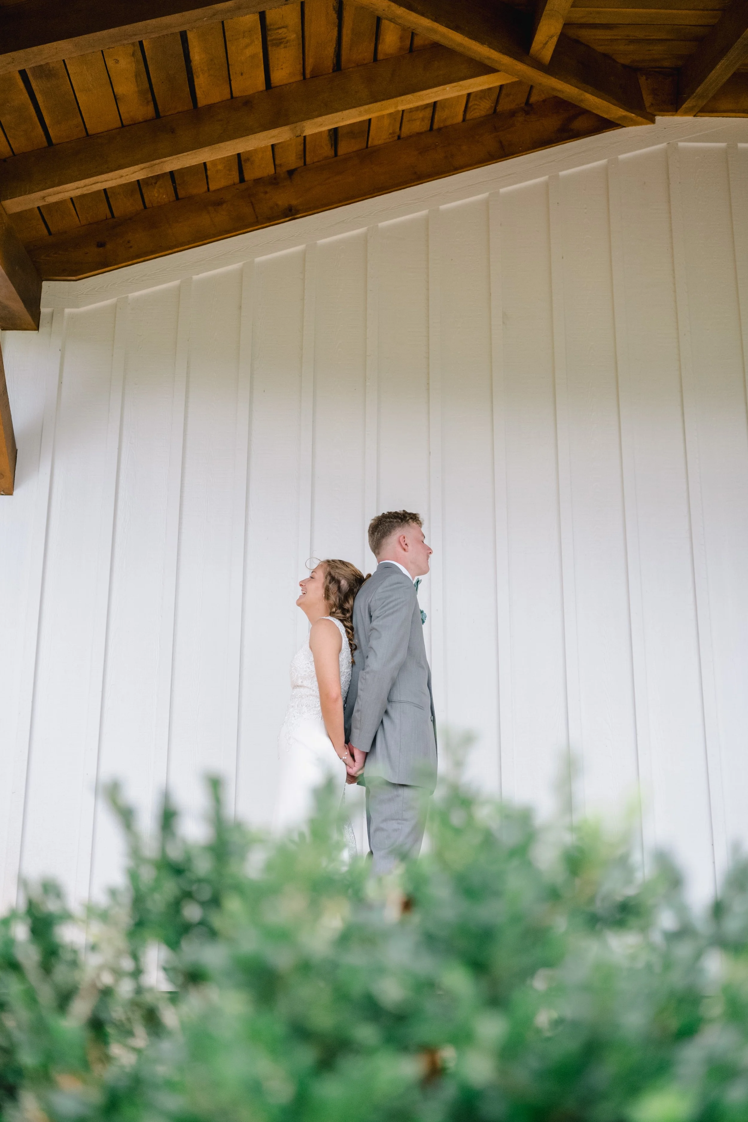 A bride and groom standing back-to-back indoors, holding hands and smiling, with a white wooden wall and wooden ceiling above them.