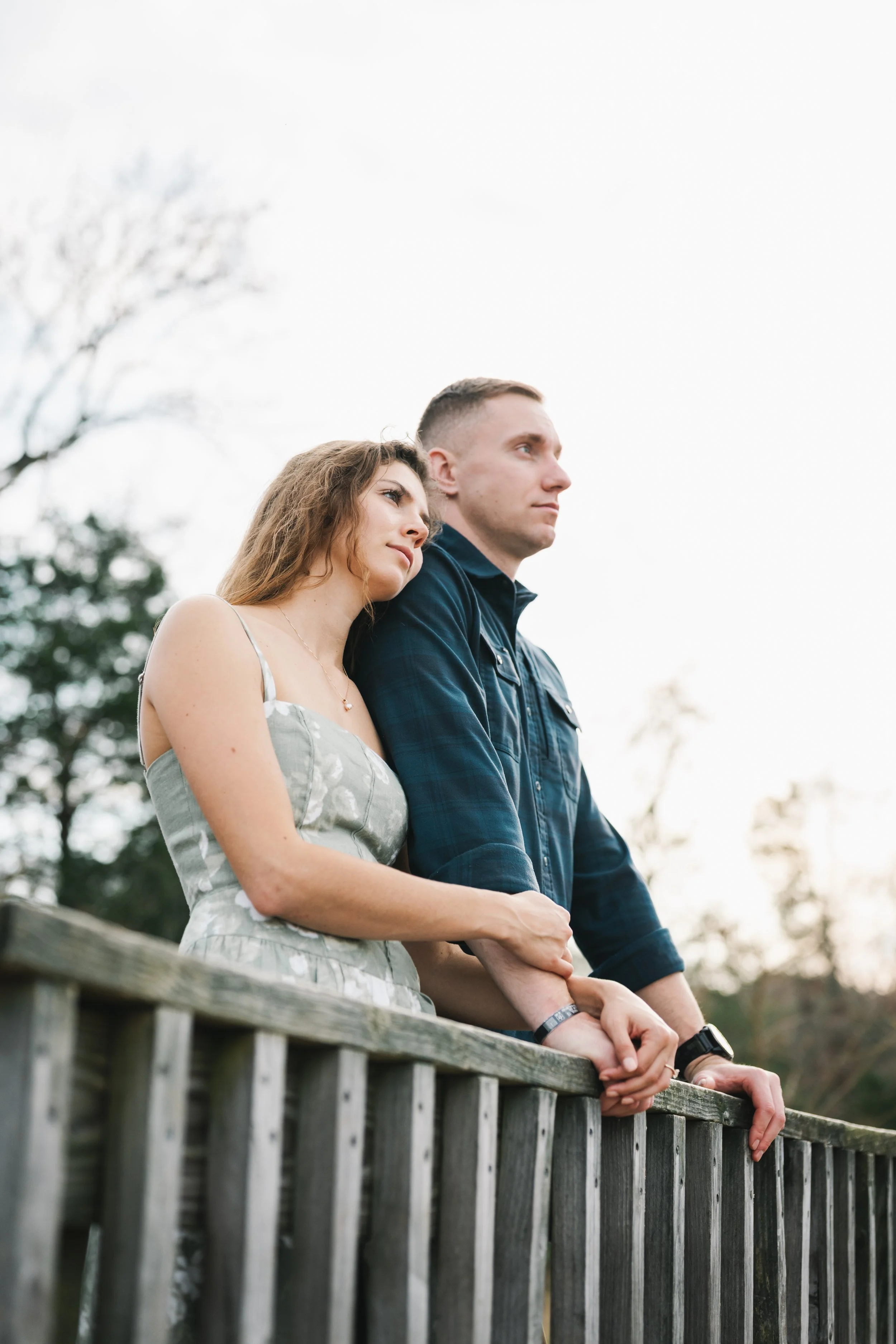 A young couple standing on a wooden bridge, holding hands and gazing thoughtfully into the distance during sunset.