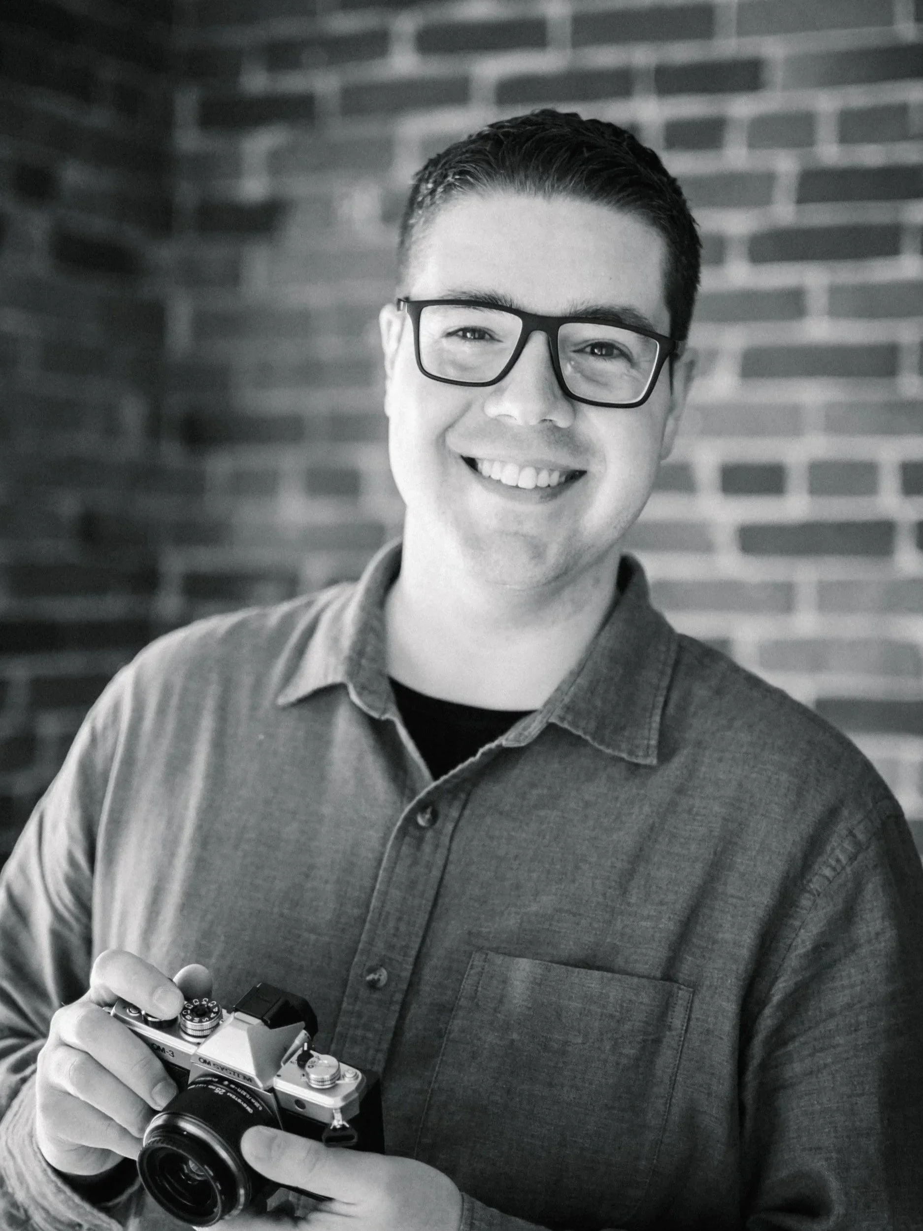 A smiling man wearing glasses holds a vintage camera, standing in front of a brick wall.