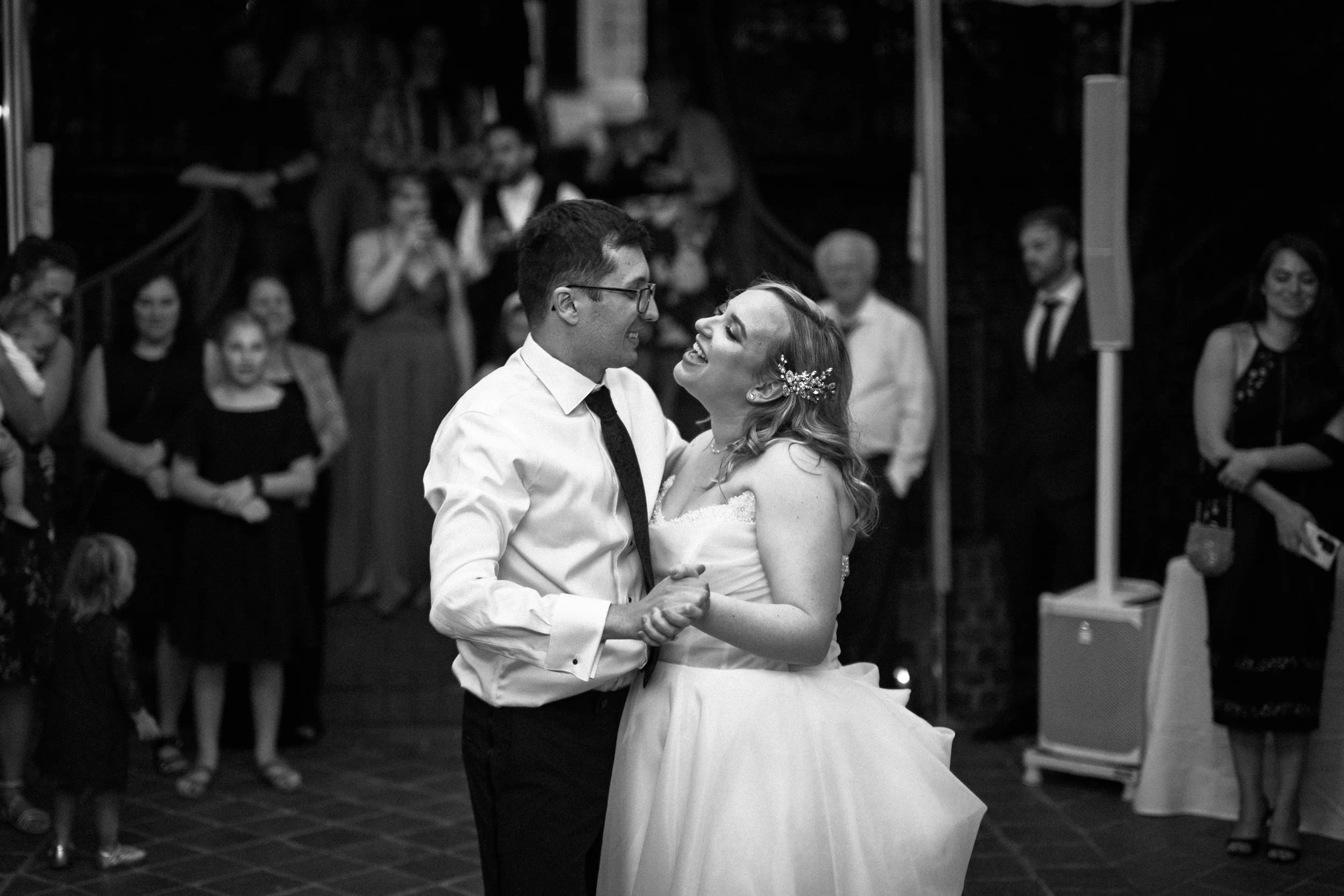 A bride and groom sharing a first dance at their wedding reception, surrounded by guests in a dimly lit indoor venue.