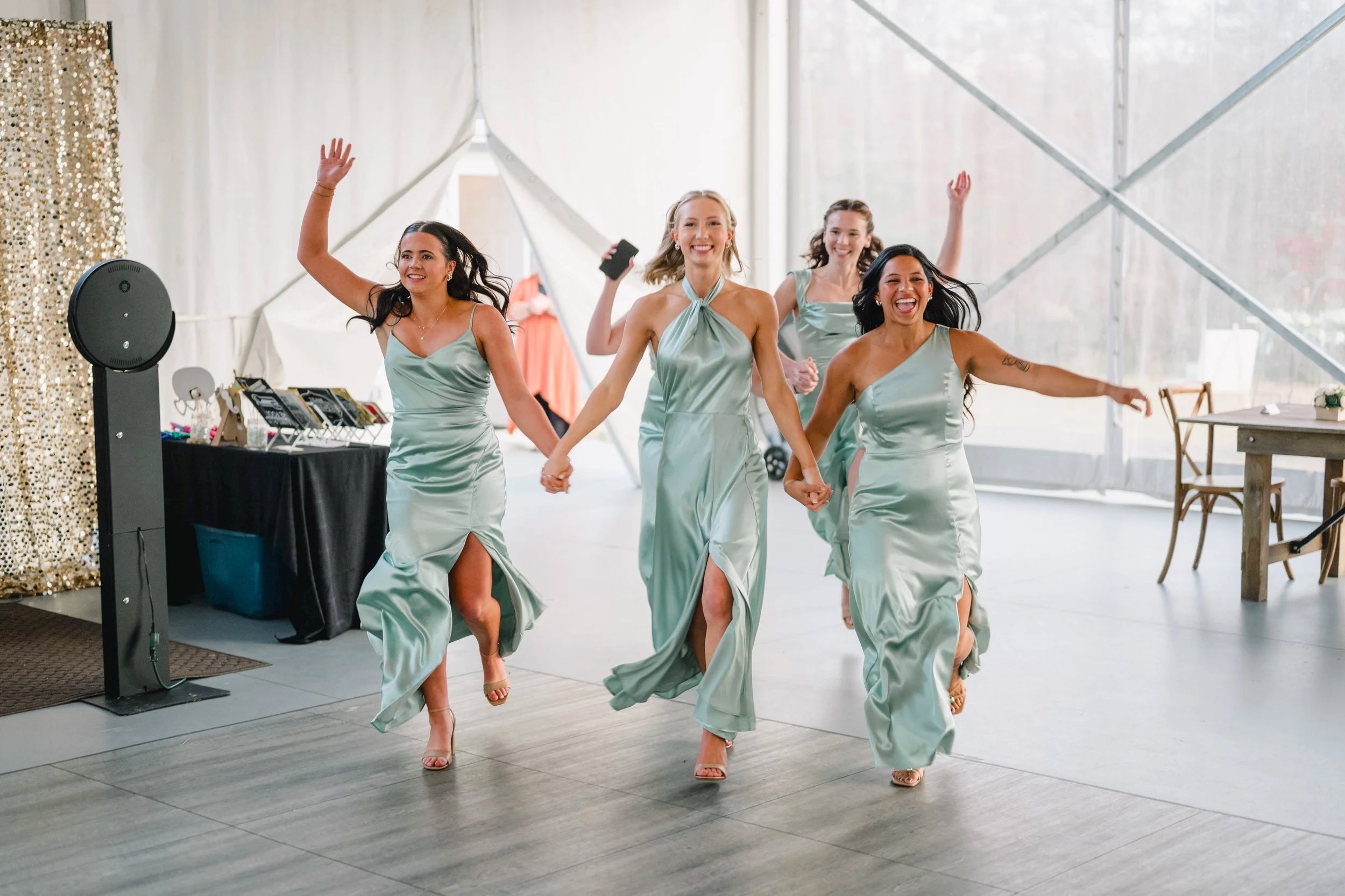 Bridal party dancing inside a reception tent, holding hands and smiling, wearing matching pastel green satin dresses.
