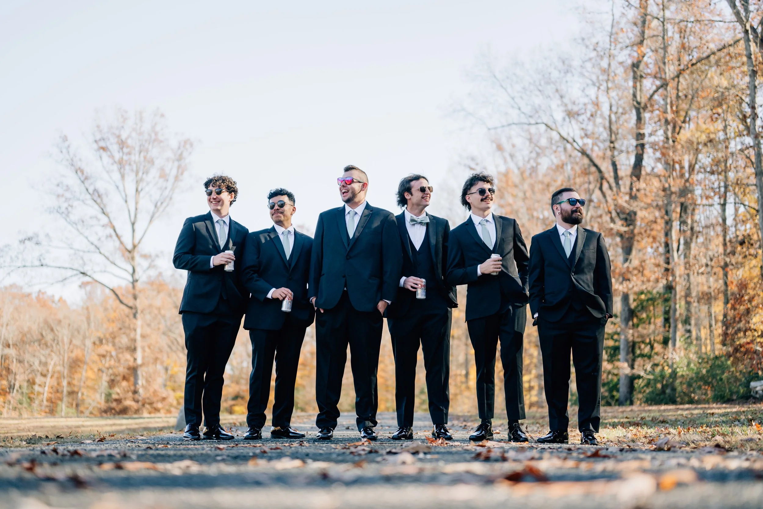 Group of men in suits and sunglasses standing outdoors in a park with Fall foliage, smiling and holding drinks.