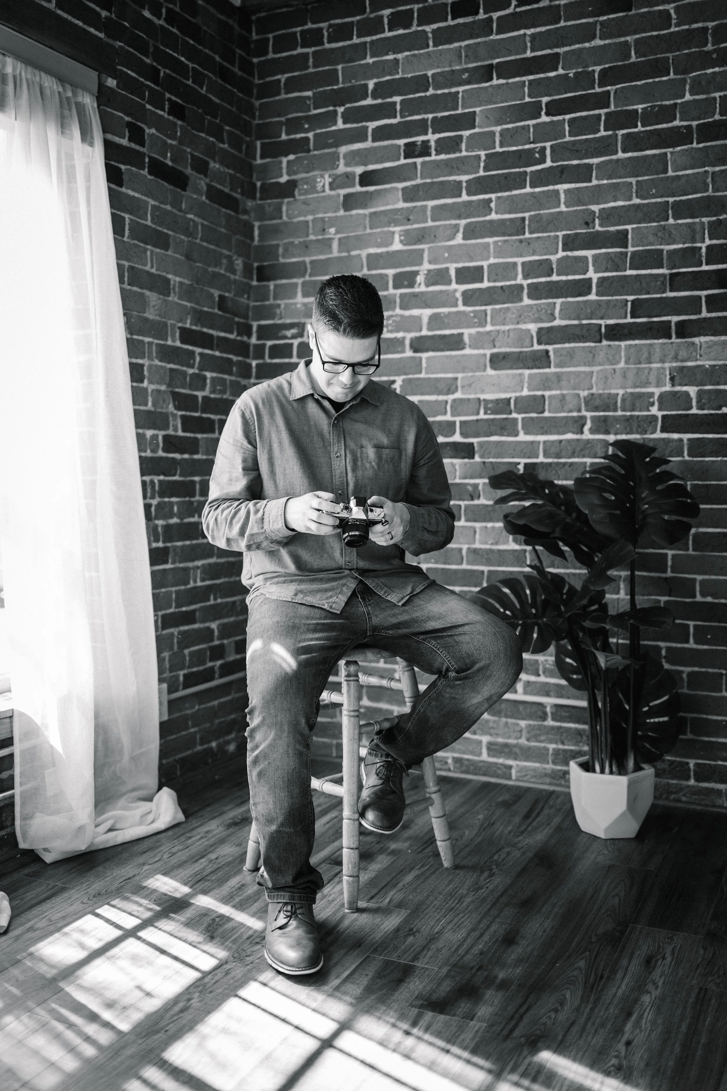 A man with glasses sitting on a wooden stool in a room with exposed brick walls, looking down at a camera in his hands, near a window with curtains and a potted plant on the floor.