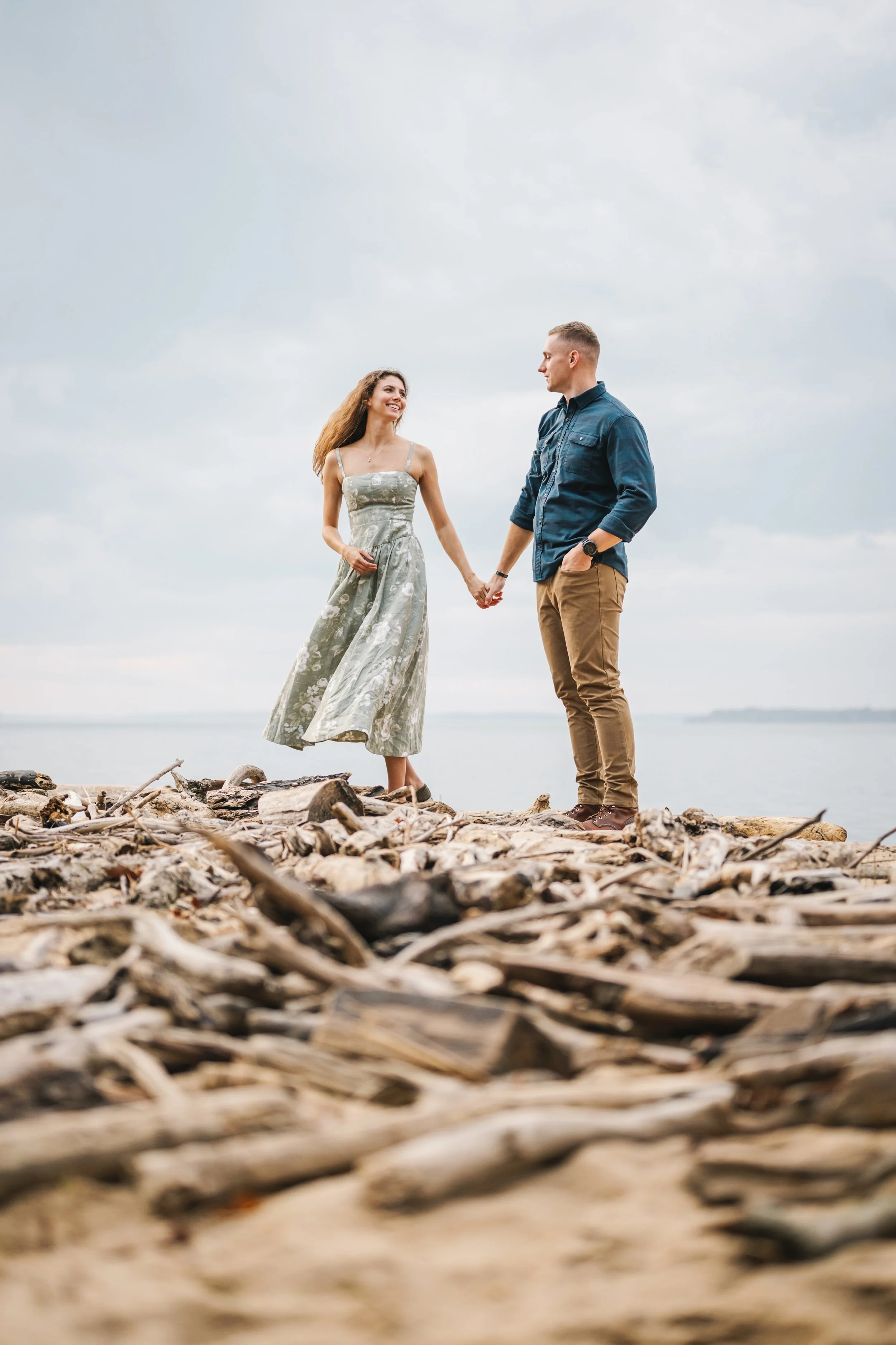 A young couple holding hands while standing on a beach with driftwood, looking at each other, under a cloudy sky.
