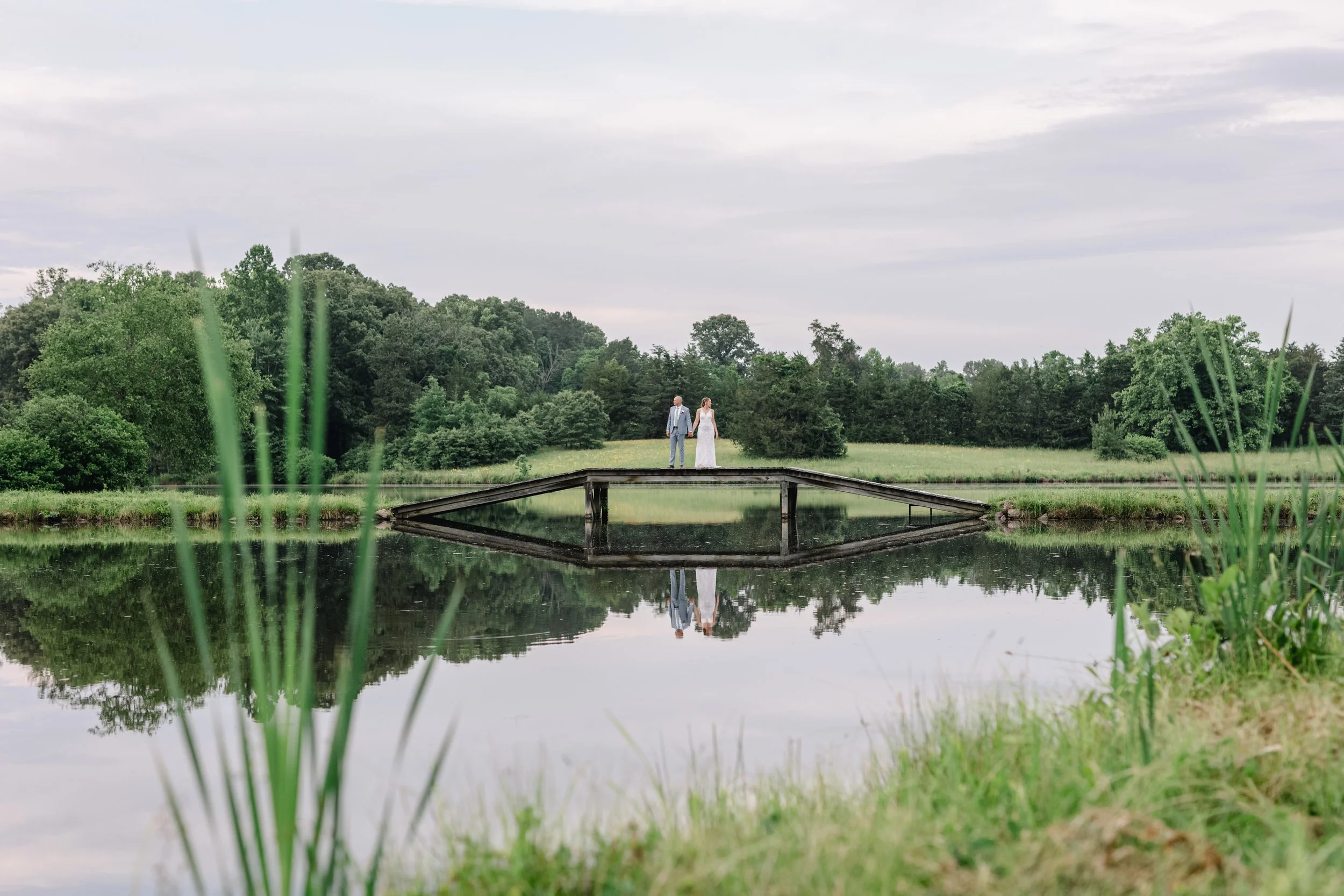 A bride and groom holding hands on a small wooden bridge over a calm pond, surrounded by green trees and grass, with a cloudy sky overhead.