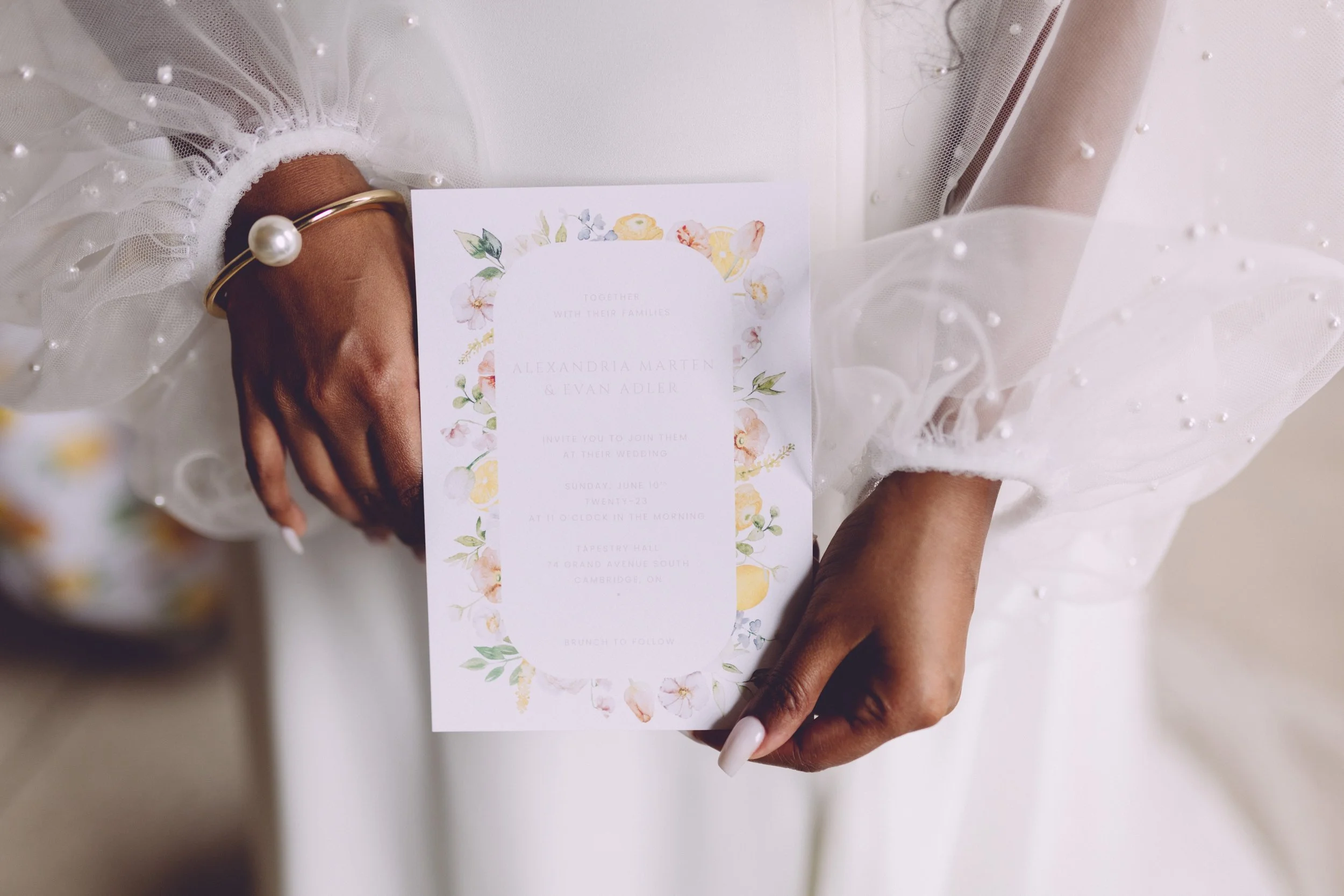 A woman holding a wedding invitation with floral design, wearing a sheer, embellished white dress and a pearl bracelet
