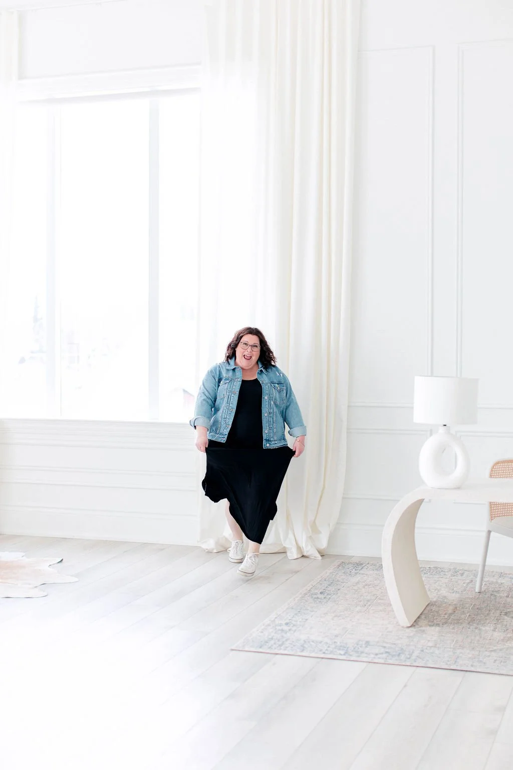 Woman with curly brown hair, glasses, wearing a denim jacket, black dress, and white sneakers standing indoors near large window with white curtains, in a bright room with white walls, hardwood floor, and modern white table with a lamp.