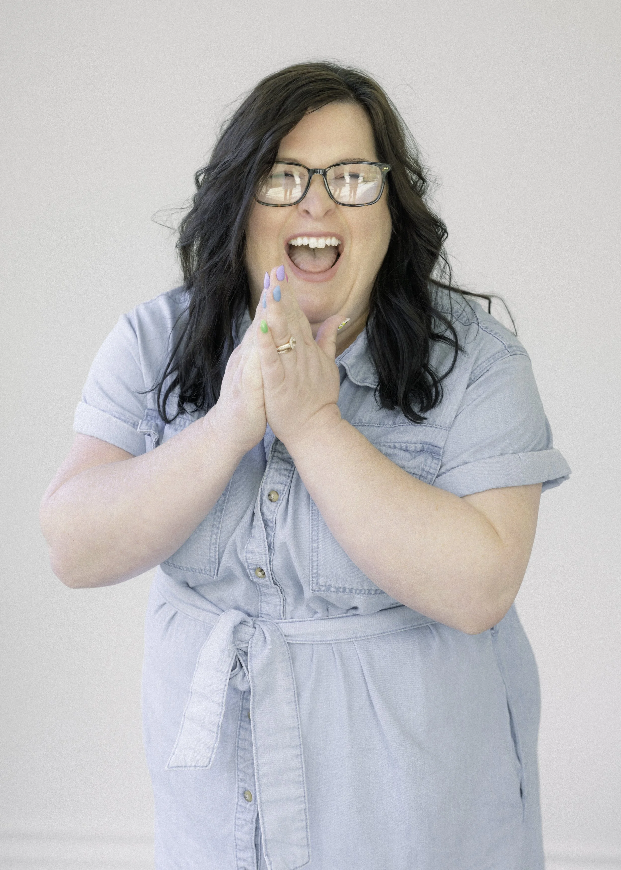 A woman with dark hair and glasses, smiling passionately with her hands clasped together in front of her face, wearing a light denim dress with rolled-up sleeves, standing against a plain light gray background.