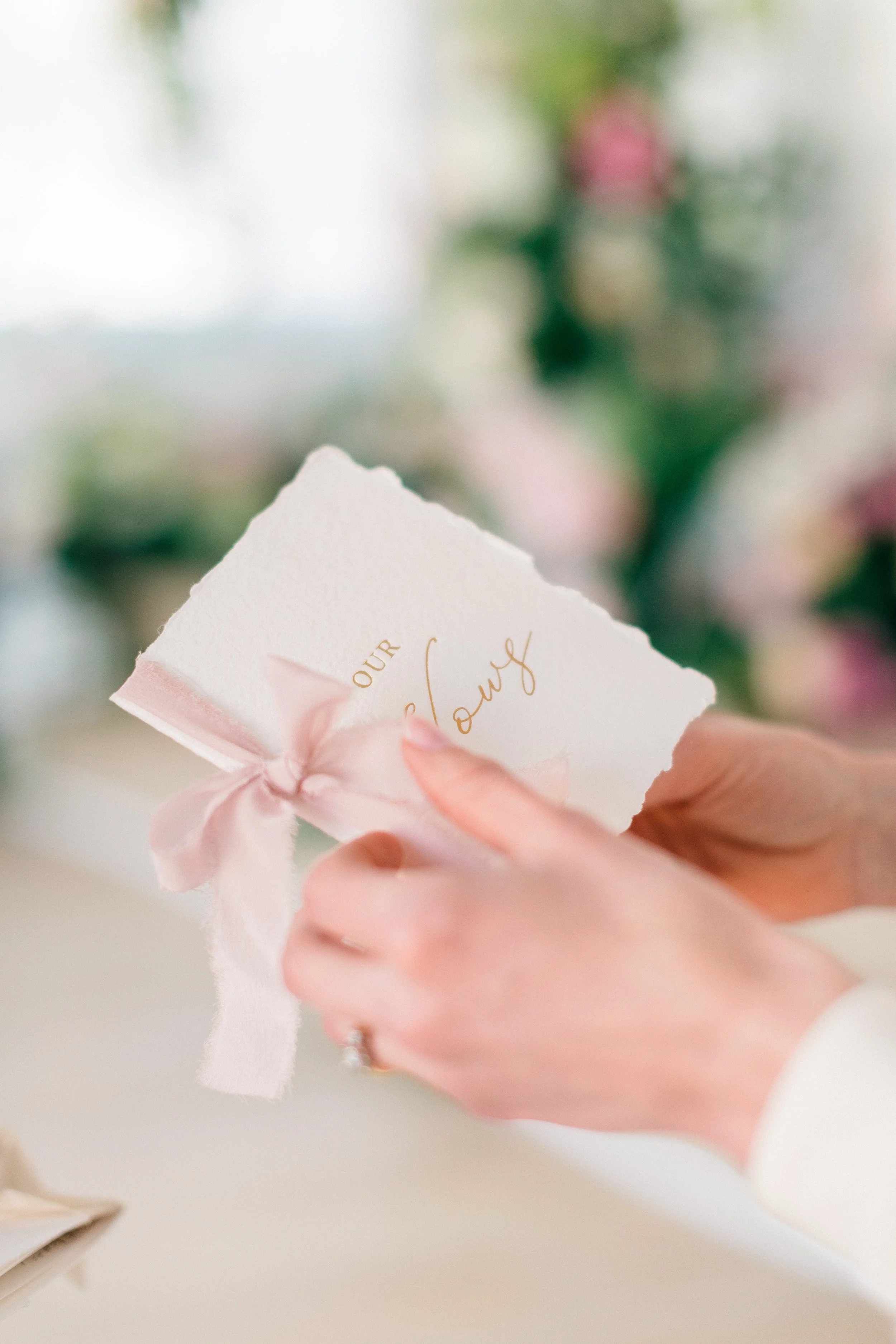 Person holding a wedding invitation with a pink ribbon, with blurred flowers in the background.