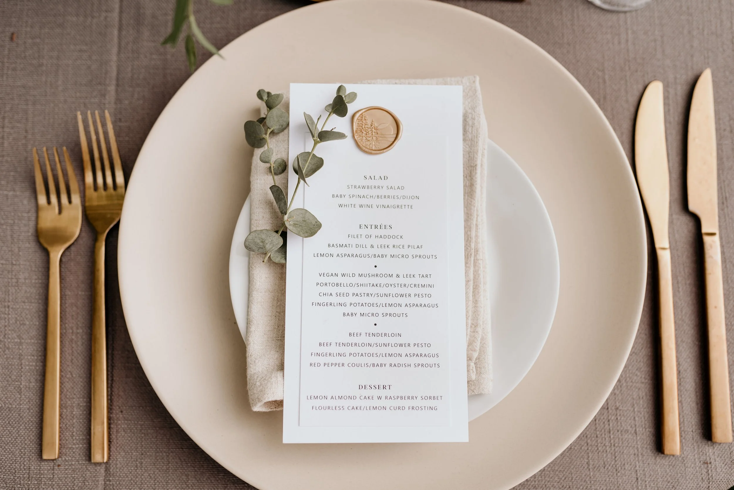 Elegant table setting with white plates, gold cutlery, a beige napkin, and a printed menu card decorated with a sprig of greenery and a wax seal, on a textured brown tablecloth.