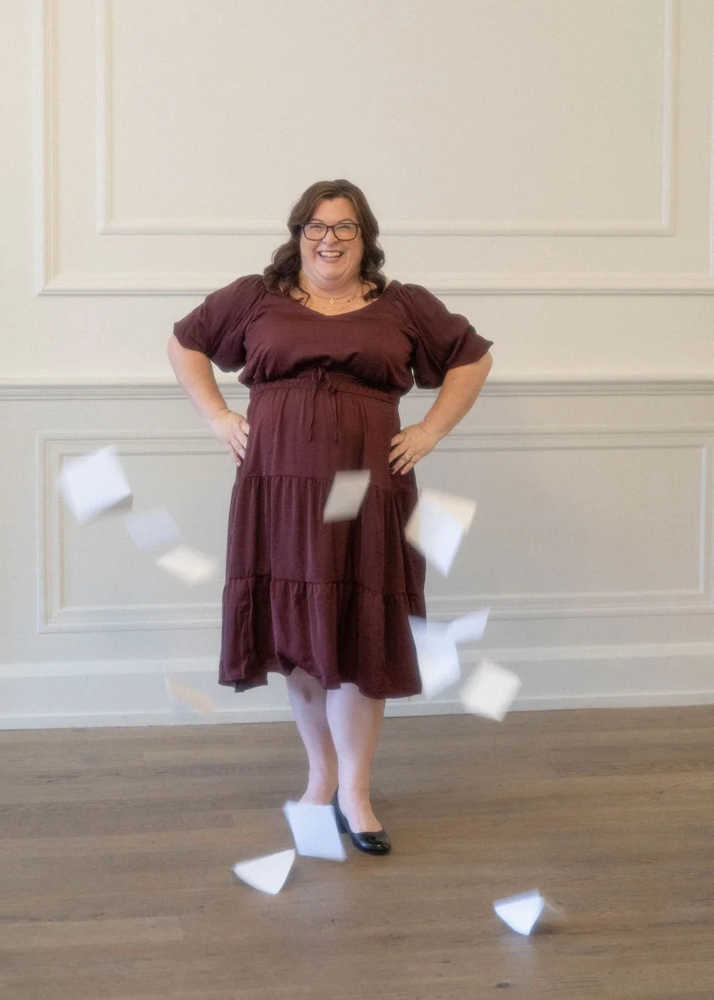 Woman with glasses smiling happy, wears a burgundy dress, standing with hands on hips, surrounded by falling white papers, in a room with beige walls and wood floor.