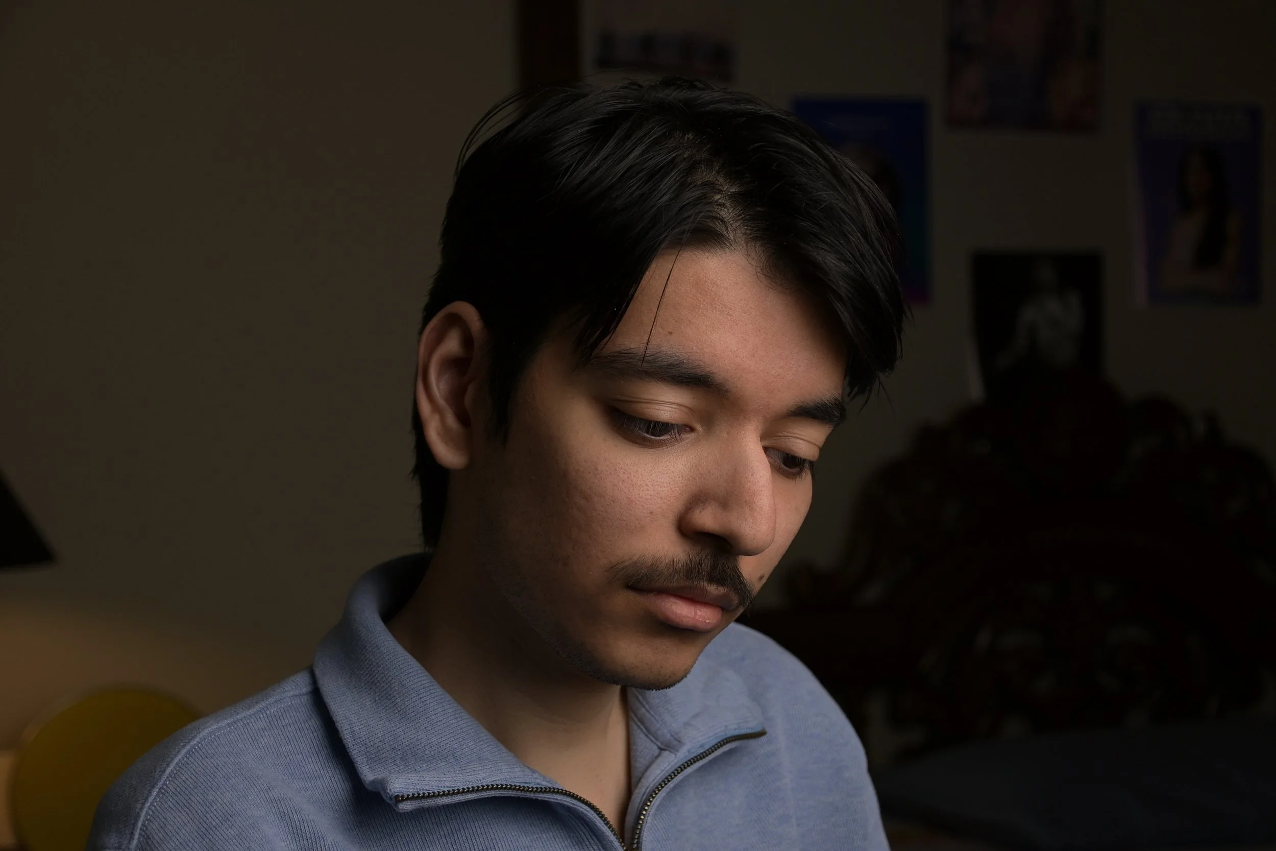 A young man with dark hair, a mustache, and a goatee looking down, wearing a gray zip-up sweater, in a dimly lit room with photos on the wall in the background.