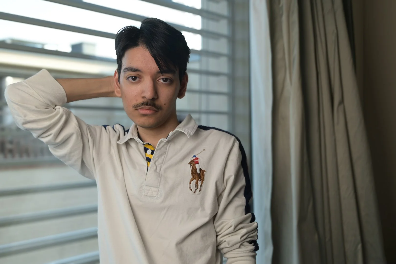 Portrait of a young man with dark hair and a mustache, standing indoors near a window with blinds and curtains. He is wearing a light-colored polo shirt with a Polo Ralph Lauren logo and has a neutral expression.