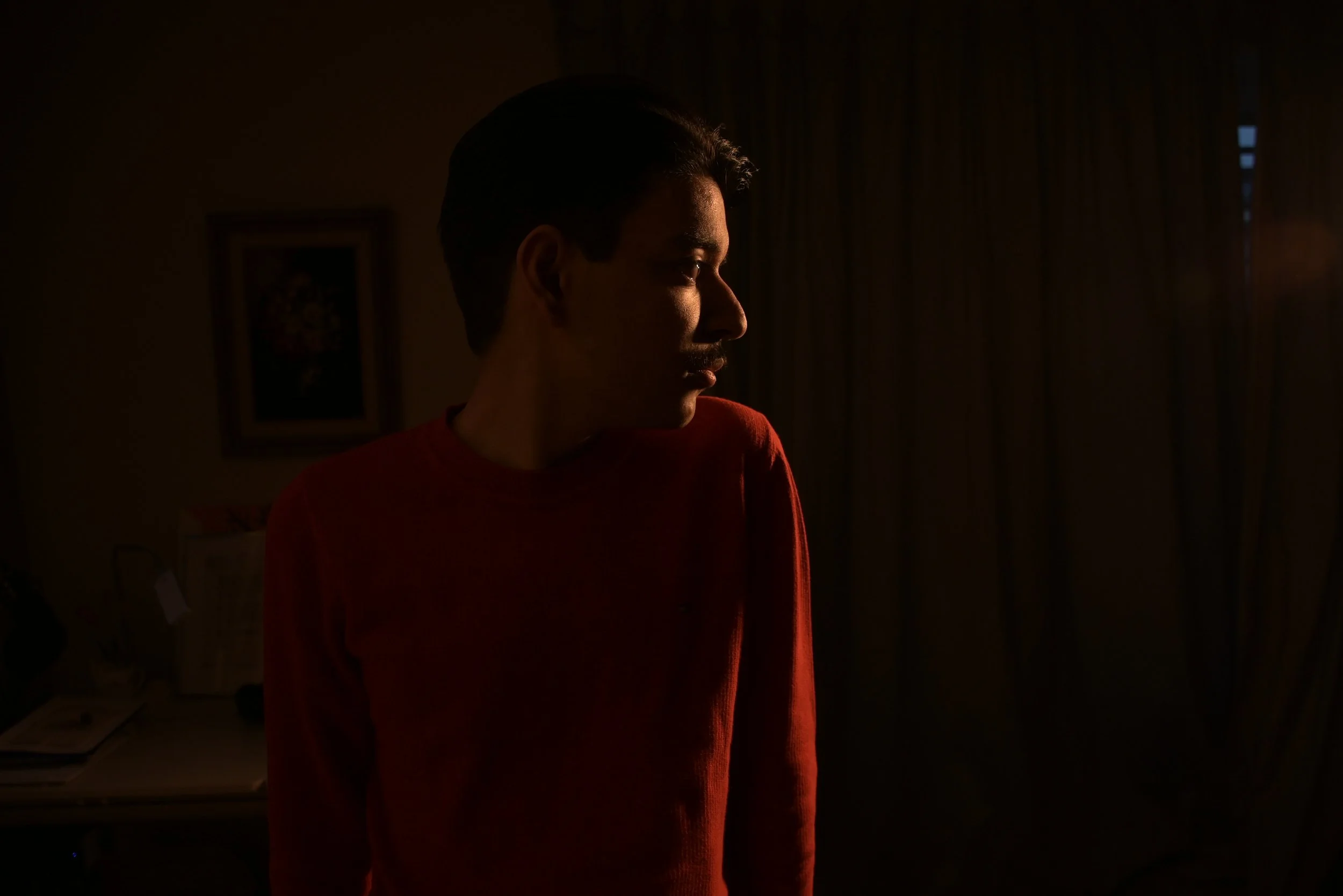 A young man with dark hair, wearing a red shirt, stands in a dimly lit room with sunlight coming through a window, casting shadows on his face in profile.