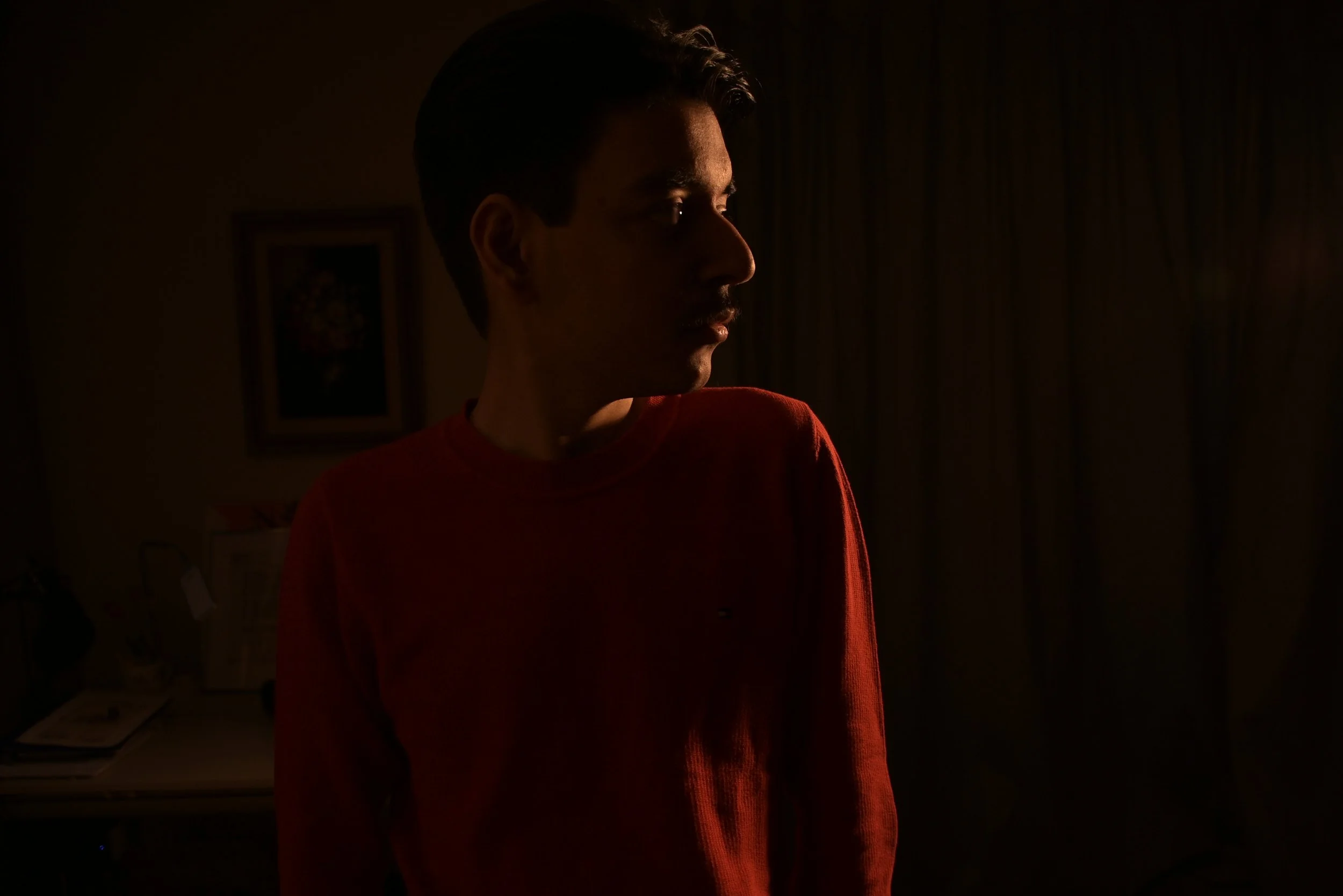 Side profile of a young man in a dark room, illuminated by a faint light on his face and shoulder, wearing a red shirt.