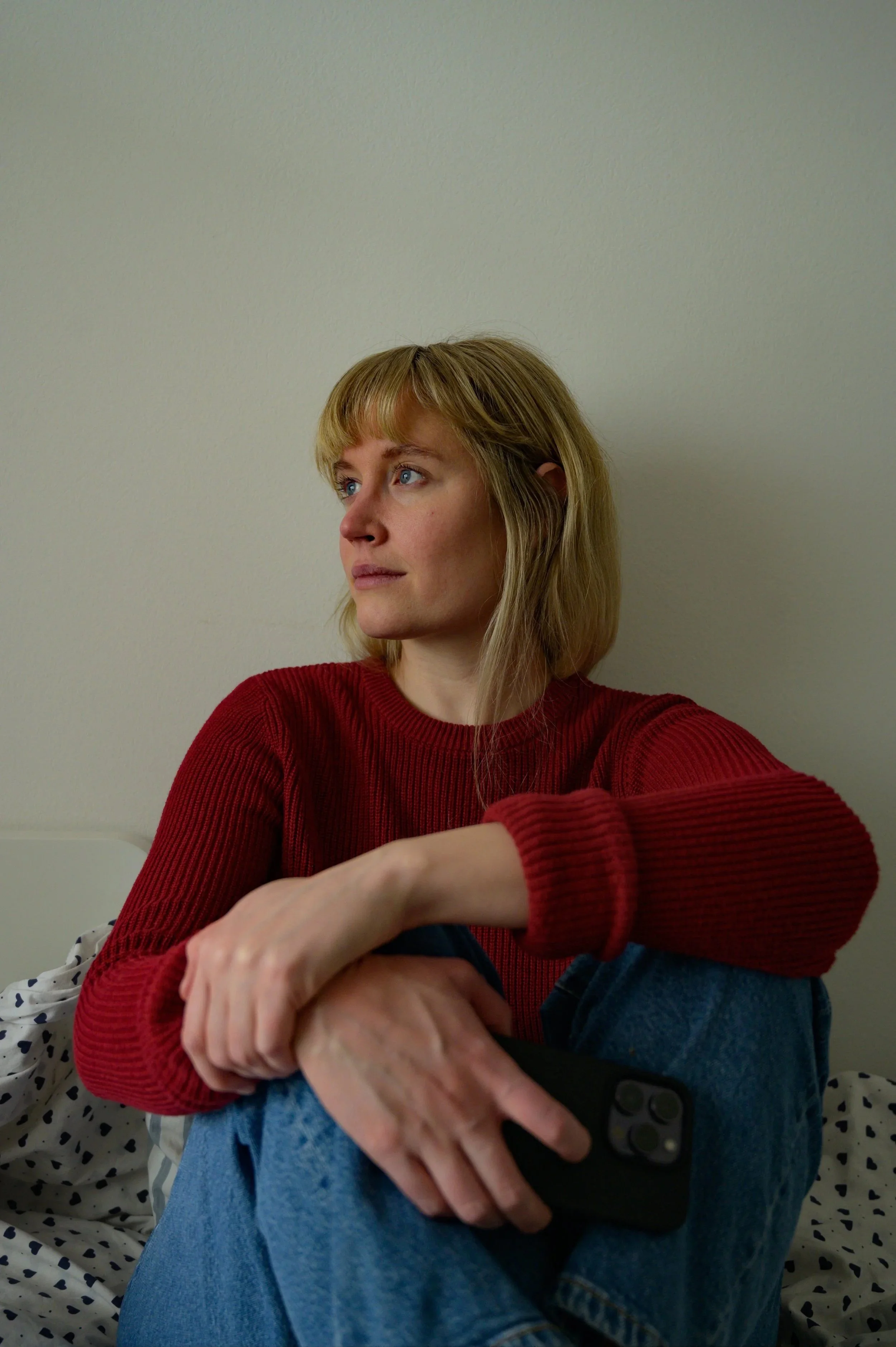 A woman with blonde hair and bangs is sitting on a bed, looking down at a smartphone held in her left hand with a gentle smile. She is wearing a textured red crewneck sweater and blue jeans. Behind her is a plain off-white wall and a white pillow wit