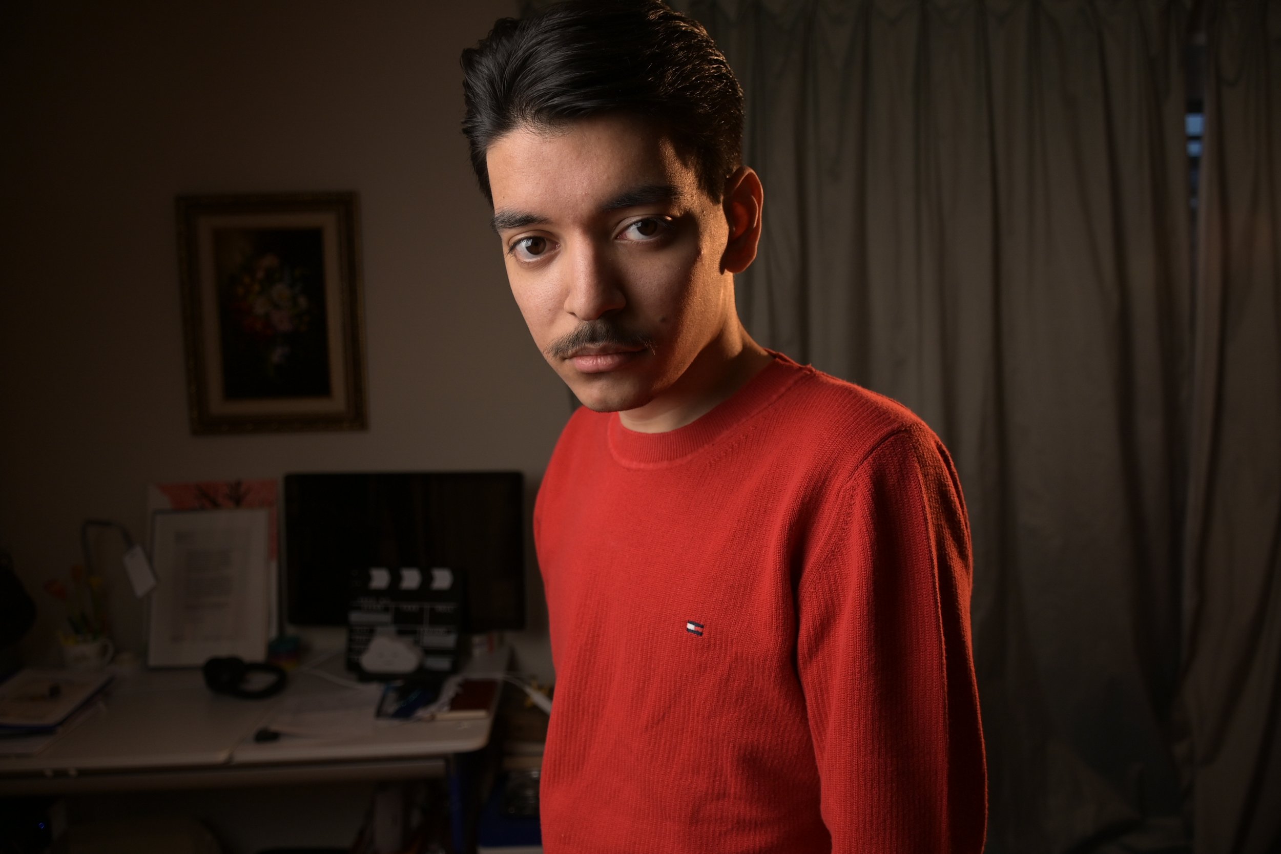 A young man with dark hair and a mustache wearing a red shirt in a dimly lit room with a desk, computer monitor, and curtains in the background.