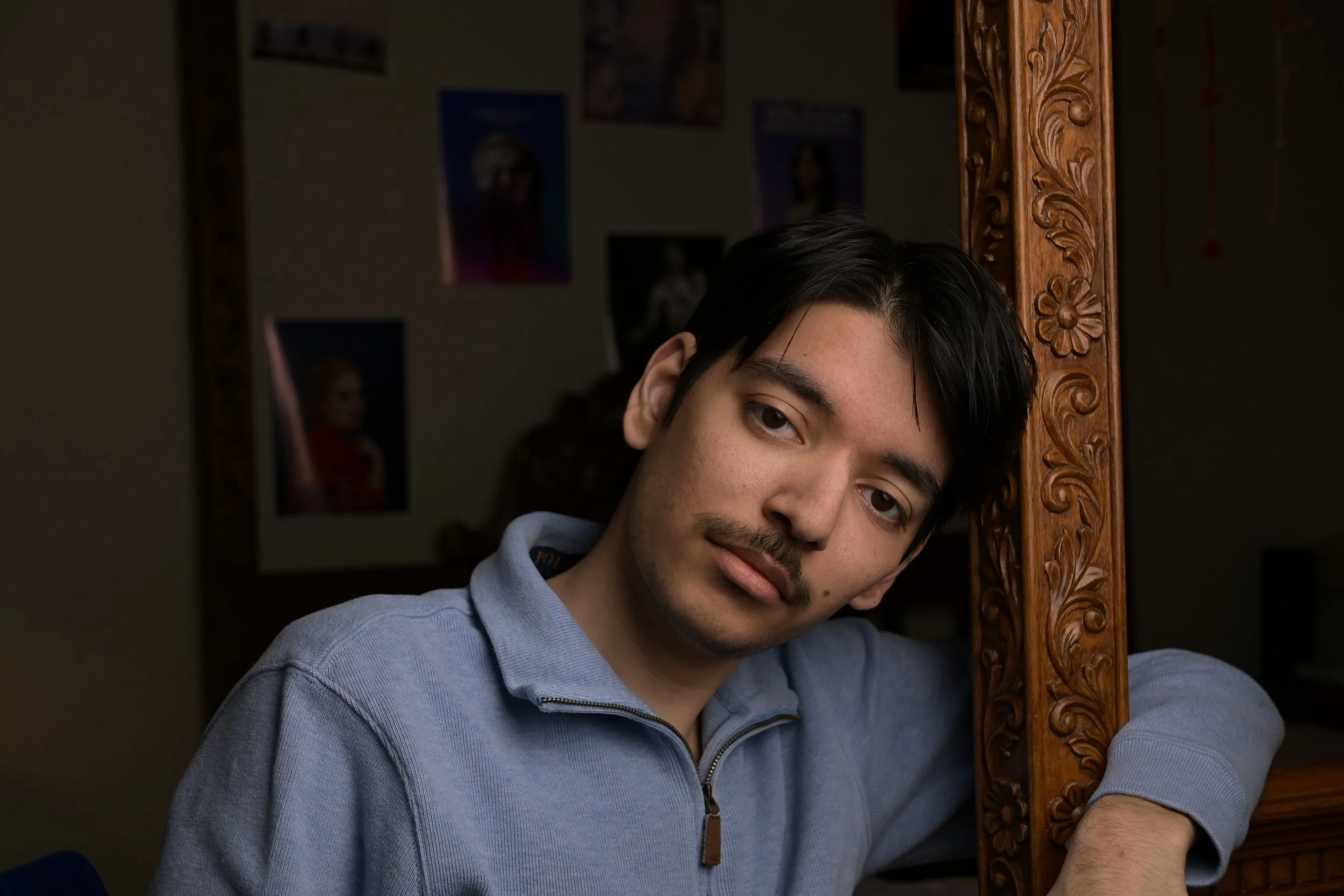 A young man with dark hair and a mustache leaning against a carved wooden frame, wearing a light blue zip-up jacket, with a softly smiling expression, in a room with dark walls and vague posters or pictures in the background.