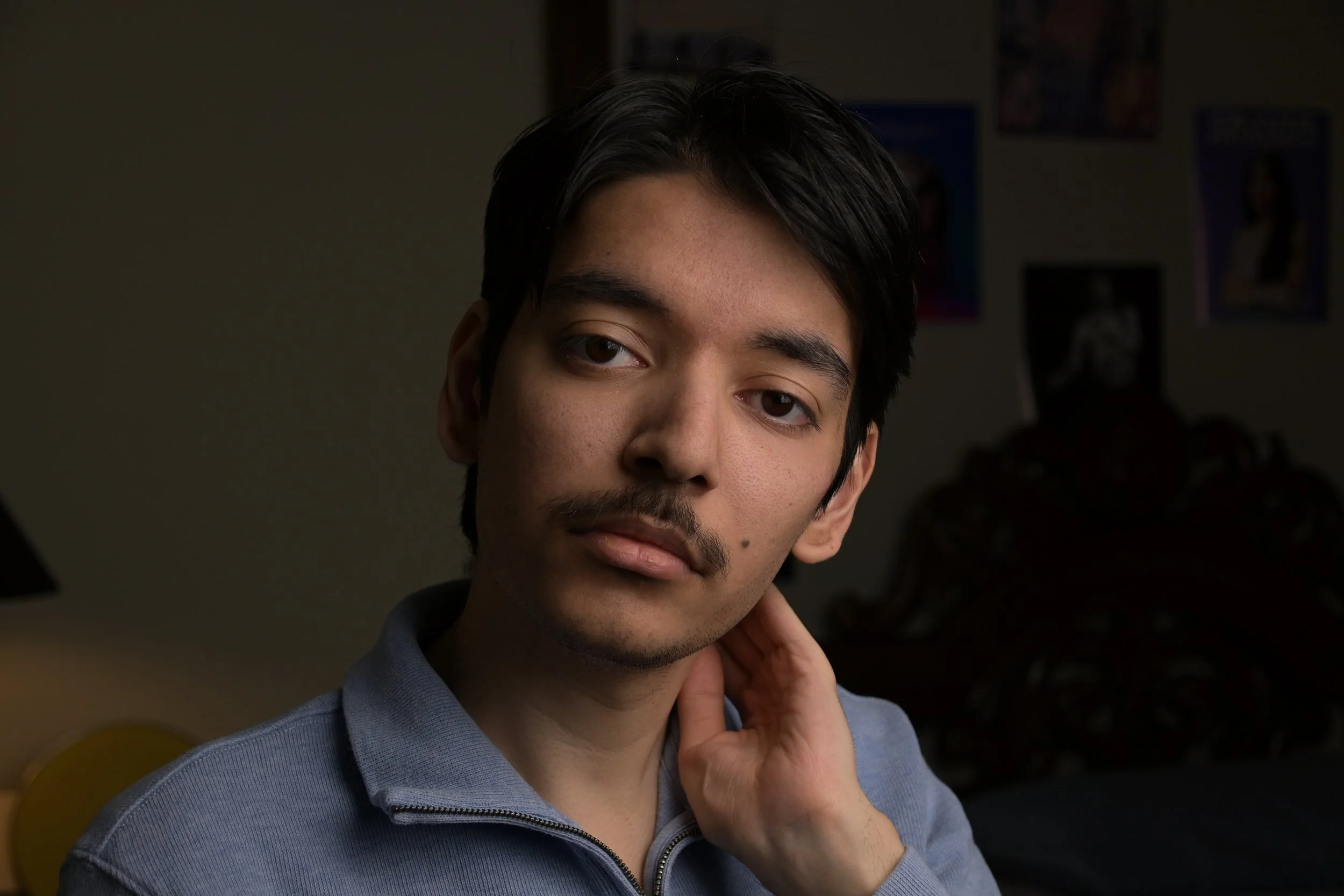 A young man with dark hair and a mustache looking at the camera, wearing a zip-up jacket, in a dimly lit room with posters on the wall in the background.