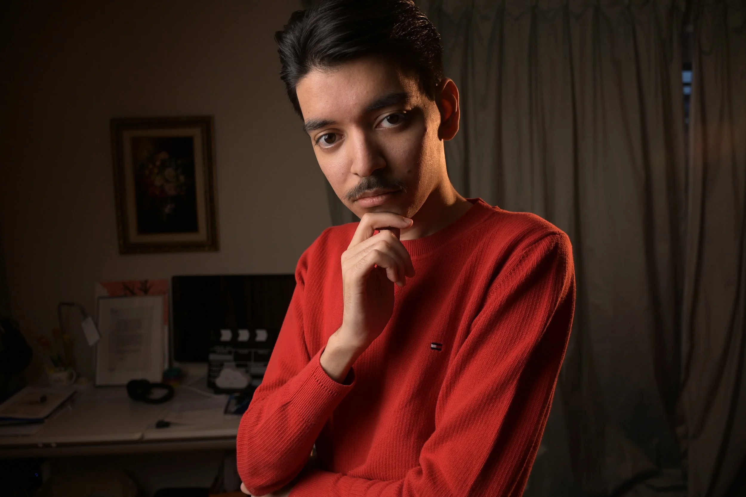 Young man with dark hair and mustache wearing a red sweater, standing indoors with a thoughtful expression, hand on chin, in front of a desk and curtains.