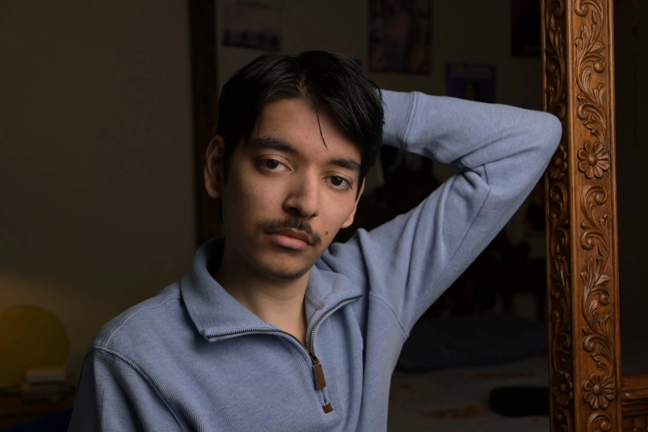 A young man with dark hair and a mustache, wearing a light blue zip-up sweater, poses with his arm resting on a carved wooden mirror frame in a dimly lit room with posters on the wall.