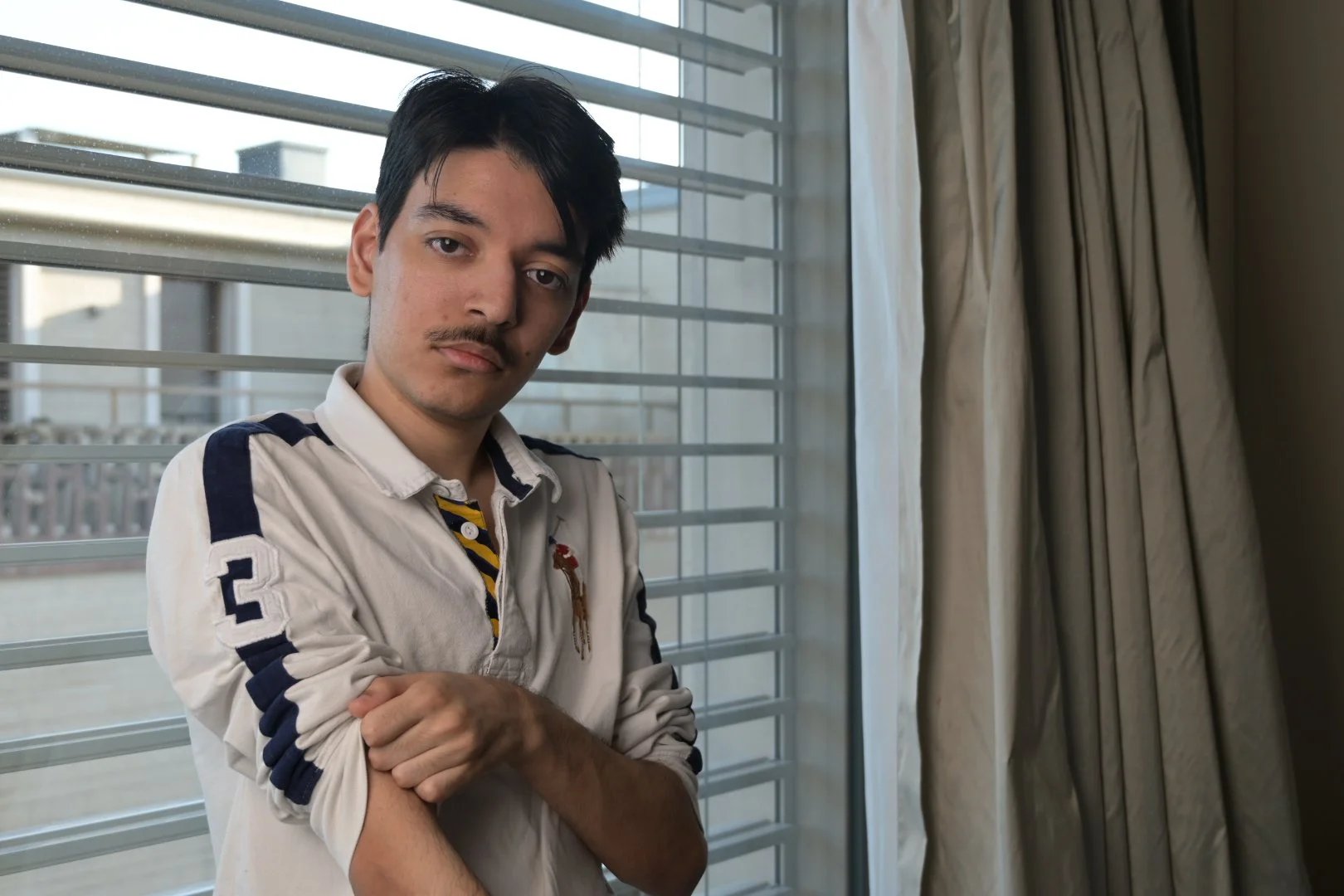 A young man with black hair and a mustache wearing a white long-sleeved shirt with navy and yellow accents, standing indoors near a window with blinds and curtains.