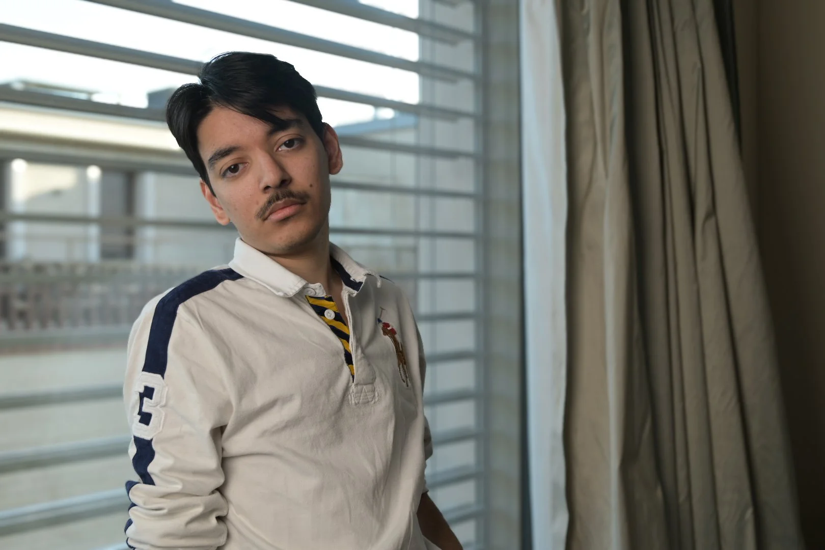 A young man with dark hair and a mustache standing indoors near a window with blinds, wearing a white collared shirt with navy blue and yellow details.