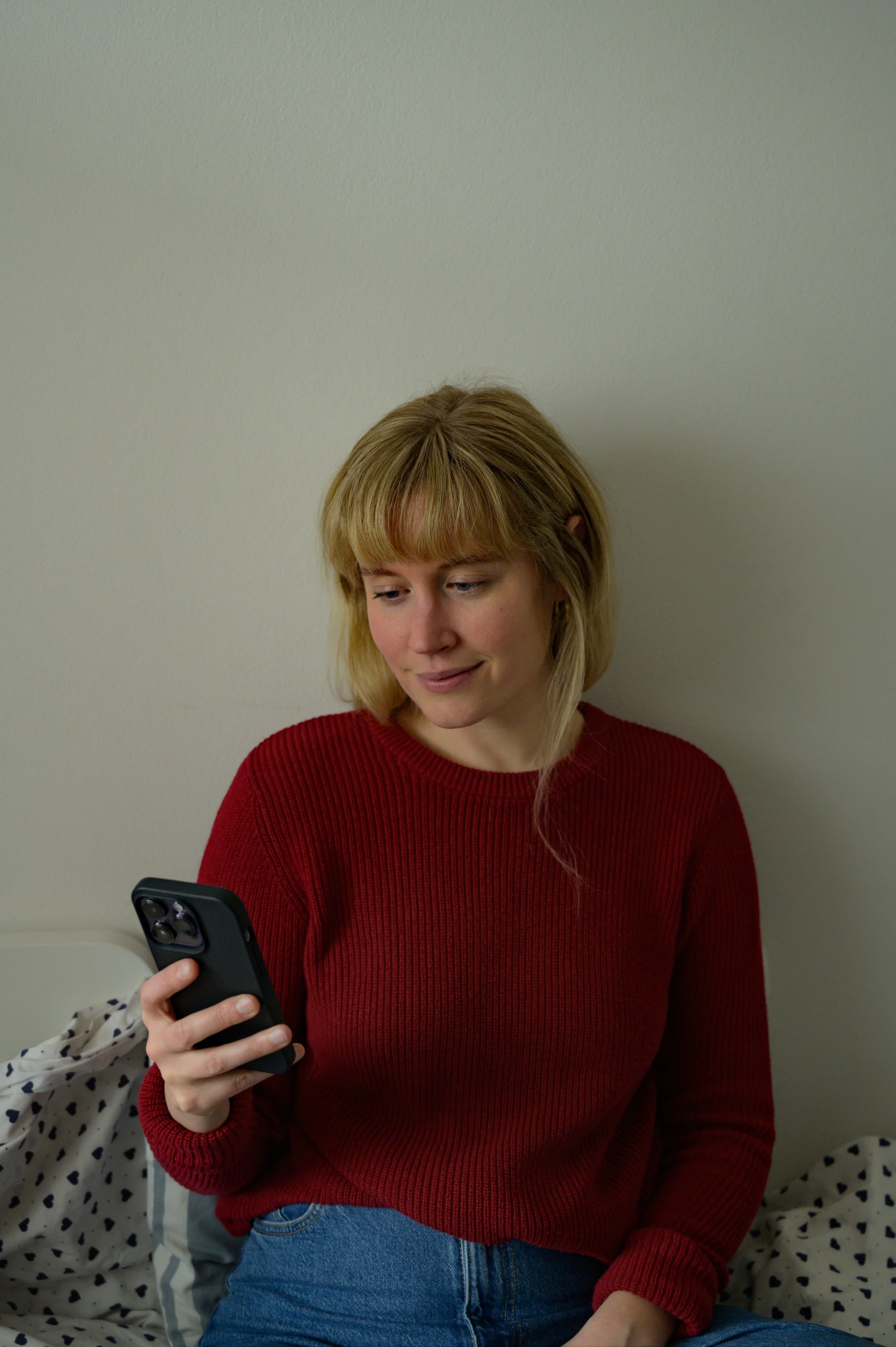 A woman with blonde hair sits on a bed, looking off to the side with a thoughtful expression. She has her arms wrapped around her knees, holding a smartphone in one hand. She is wearing a red ribbed sweater and blue denim jeans. The background is a s